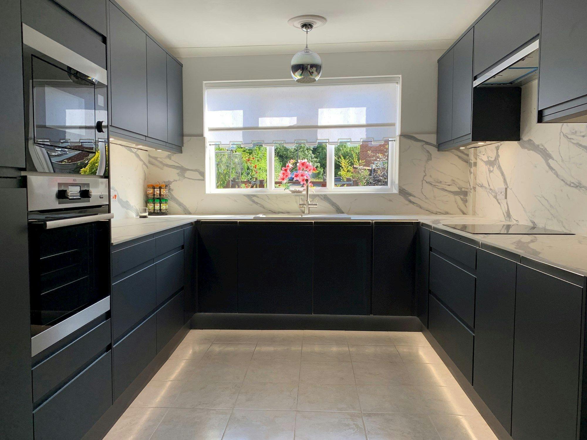 A modern kitchen with sleek black cabinetry and marble countertops, featuring integrated appliances and a central sink. Natural light illuminates the space through a large window with garden views.