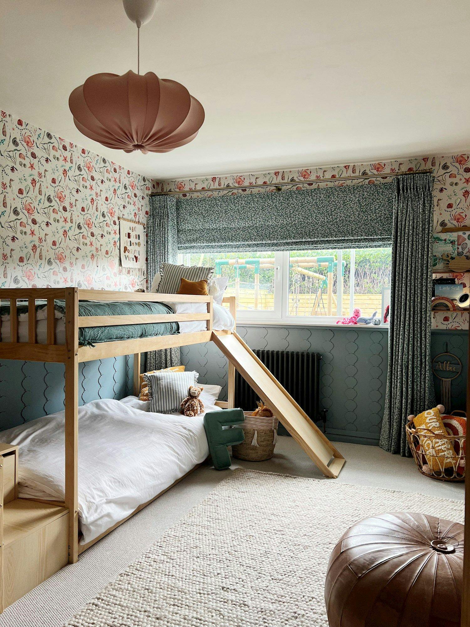 A wooden bunk bed with a slide and green bedding is in a children's room with floral wallpaper. Toys are scattered, and sunlight streams through a large window.