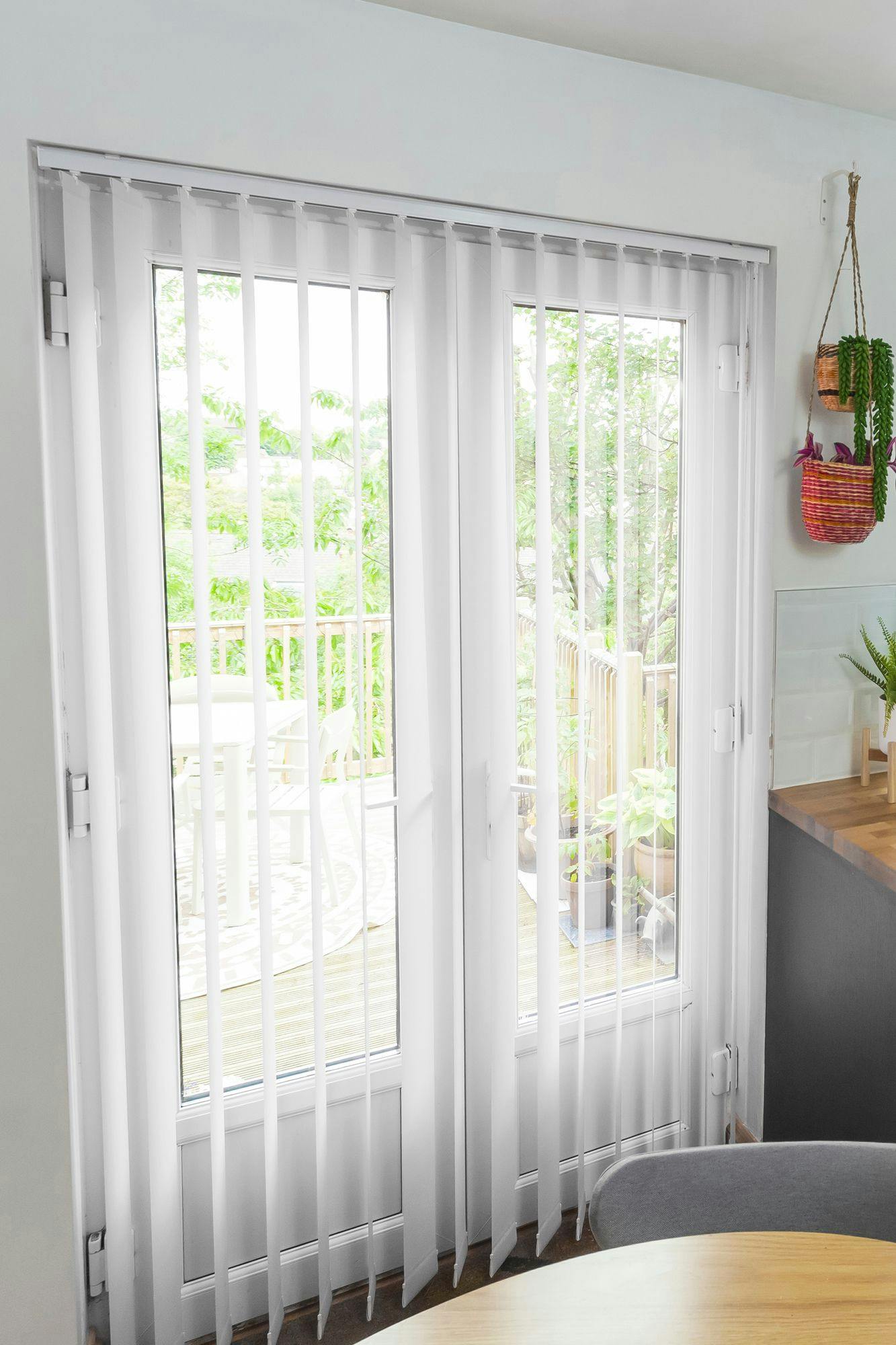 Vertical blinds cover glass doors, partially open to reveal a sunny outdoor patio with greenery. Indoors, a wooden counter and hanging baskets with plants add warmth and color.