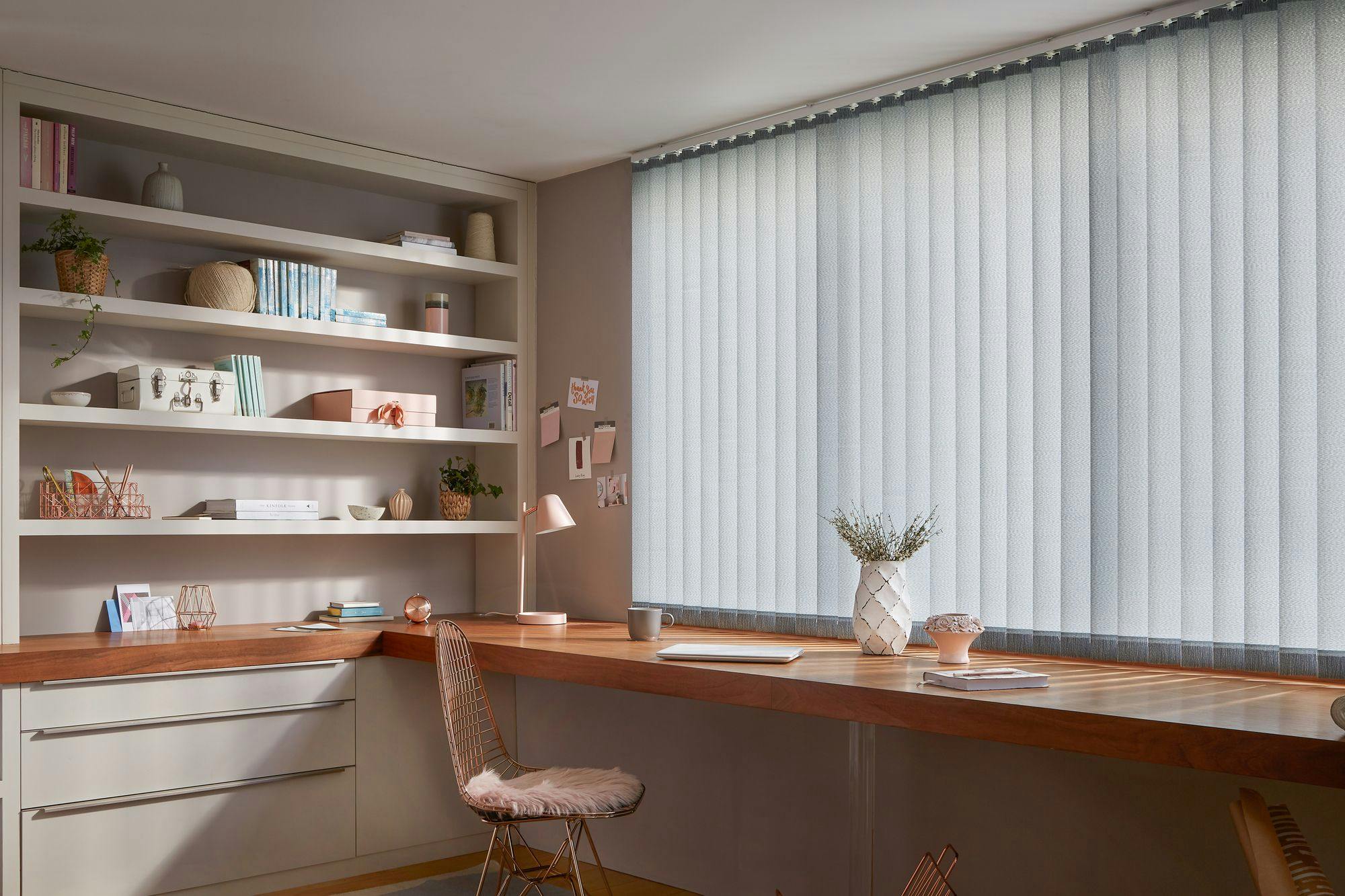 Desk with a wooden surface holds a lamp, laptop, and vase with dried flowers. Nearby bookshelves display books, decorative items, and plants. Vertical blinds cover a large window.