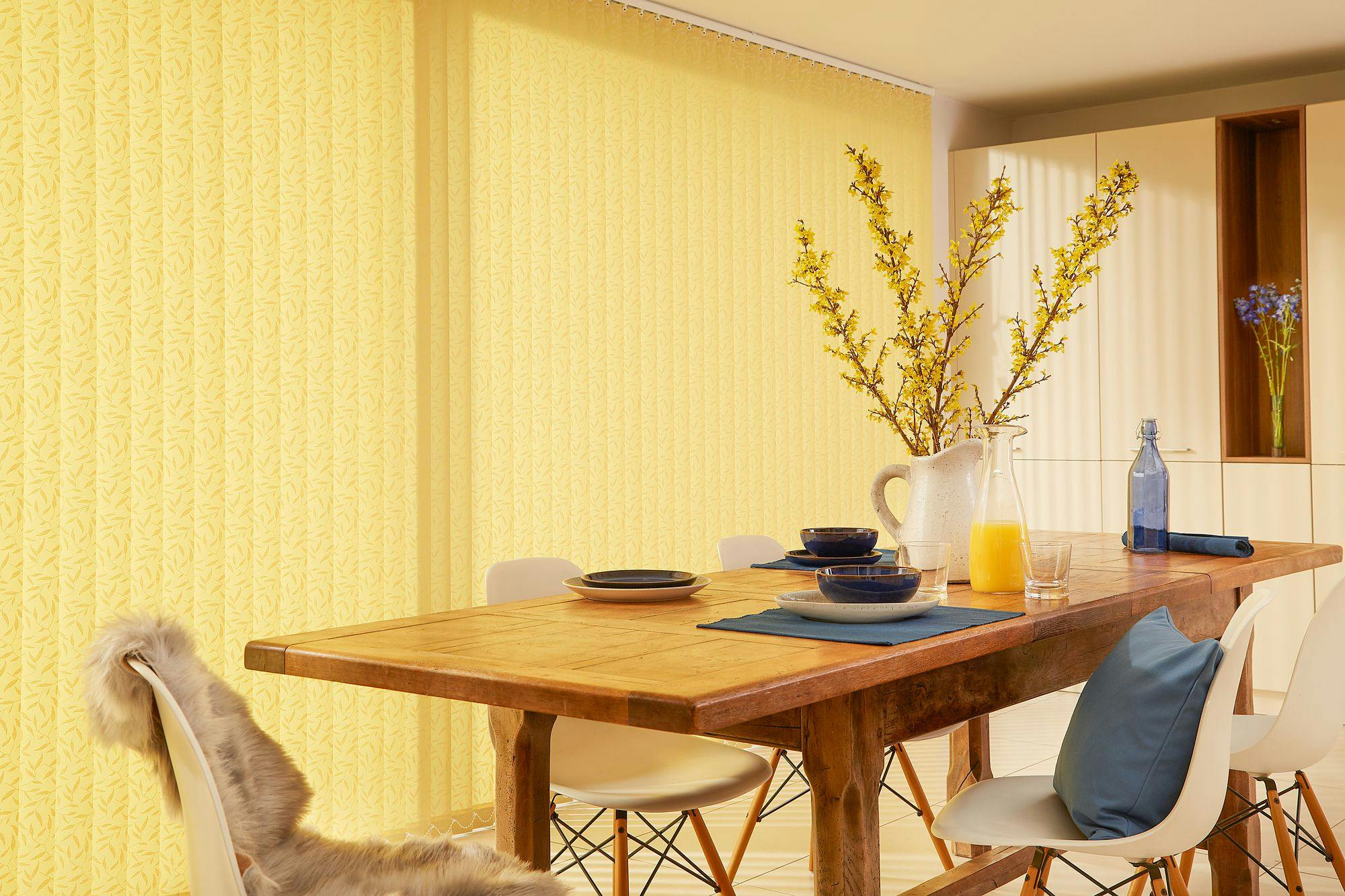 A wooden dining table set with blue dishes and orange juice sits under a large jug of yellow flowers, surrounded by white chairs against yellow-patterned vertical blinds in a bright kitchen.