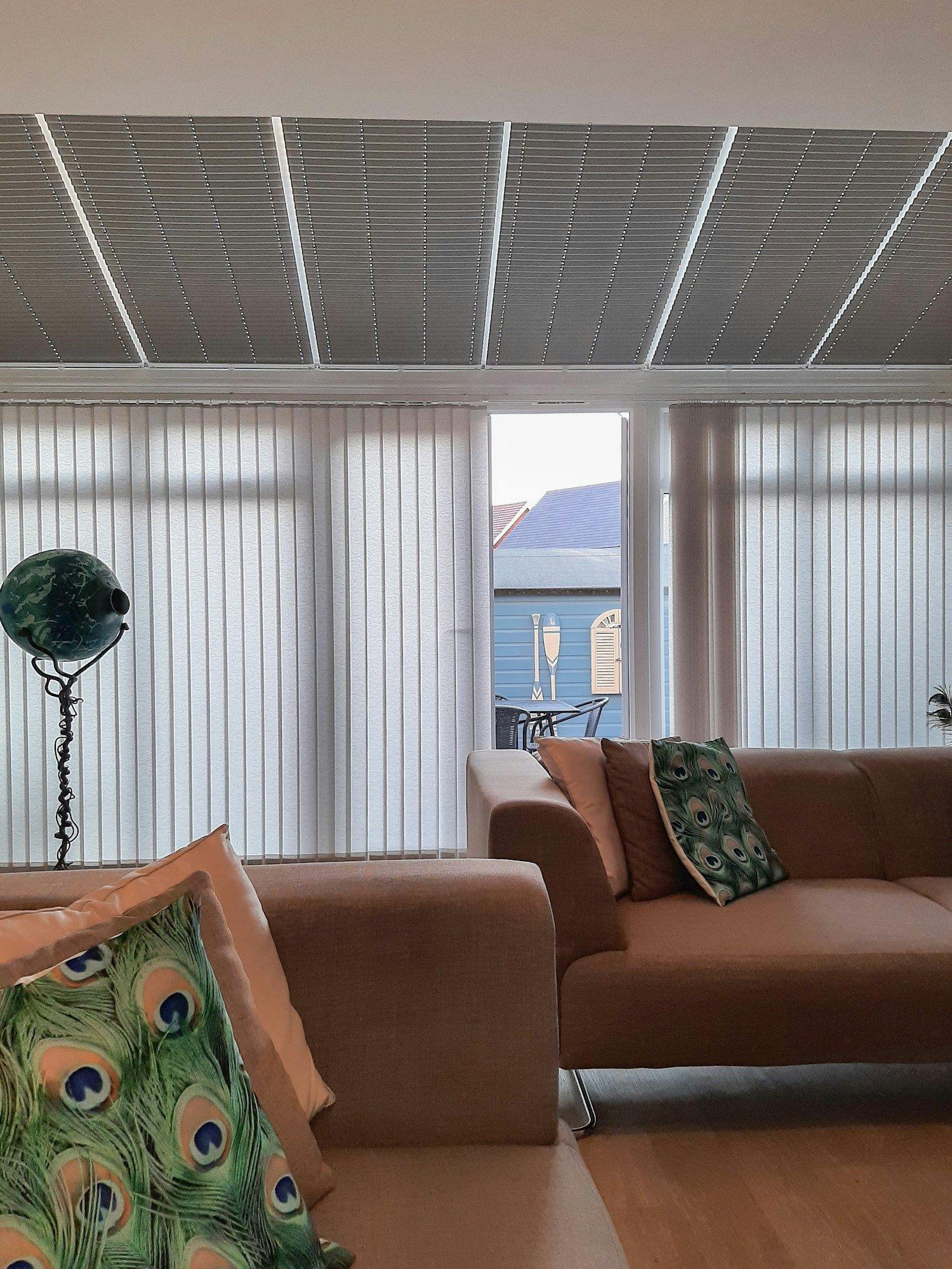 Brown sofas with peacock-patterned cushions sit in a living room, facing floor-to-ceiling windows with vertical blinds. Outside, blue buildings are visible, and a globe lamp stands nearby.
