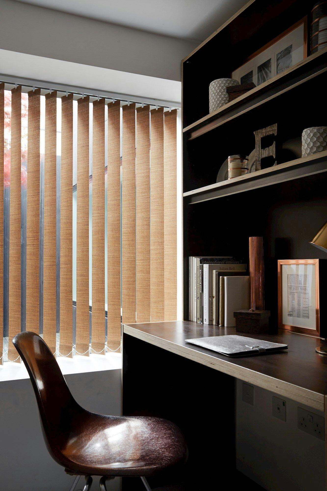 A brown chair stands near a desk with a closed laptop and bookshelves. Vertical blinds cover the window, casting light onto the modern, neatly organized workspace.