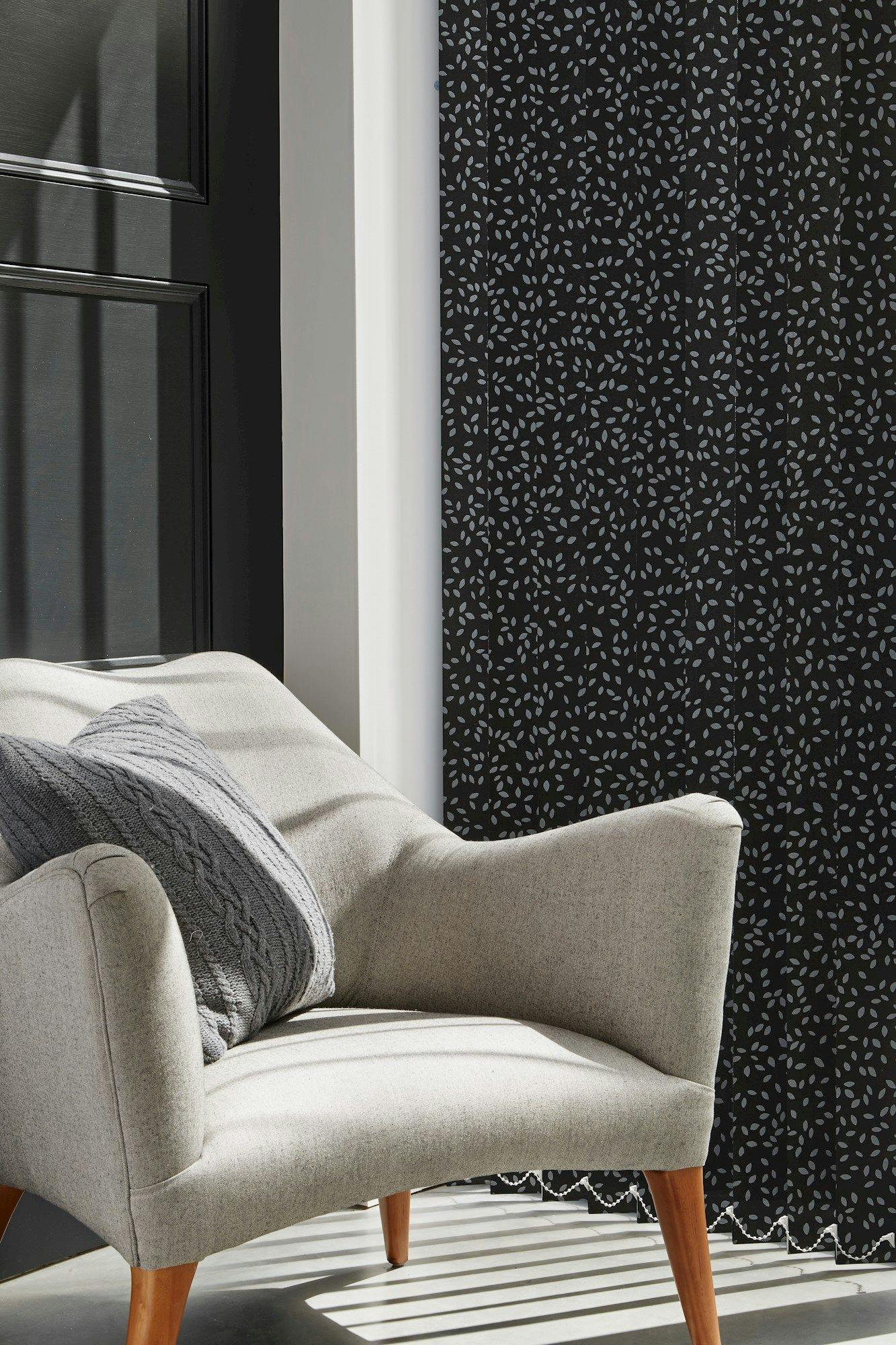 A beige armchair positioned against a wall with a black curtain featuring a white leaf pattern. A gray knitted pillow rests on the chair, with sunlight casting shadows.