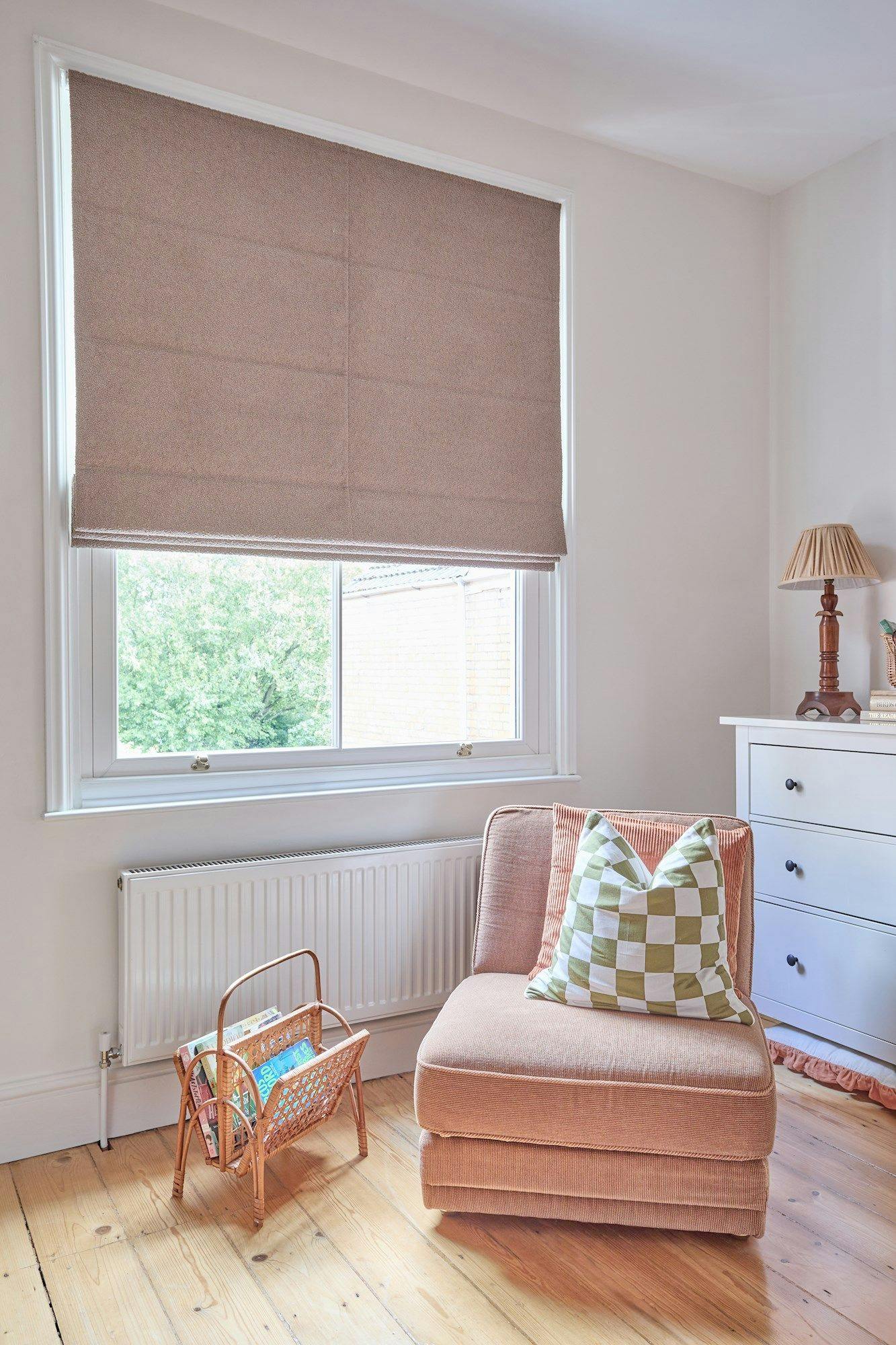 A brown Roman shade covers a window. Below, a cushioned chair holds a green-checkered pillow. Nearby, a wicker magazine holder rests on wooden flooring, beside a white dresser with a lamp.