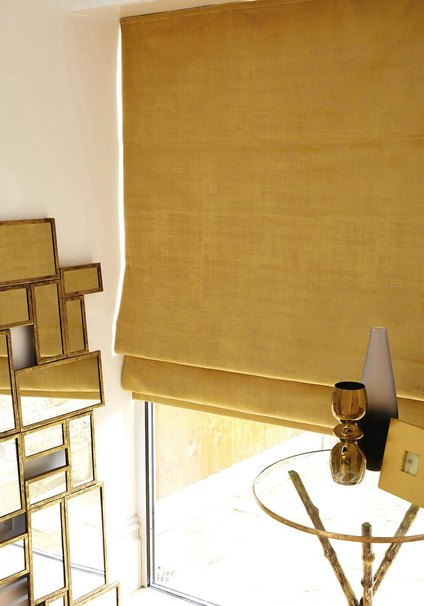 A gold fabric blind covers a window, next to a geometric mirror on the wall. A glass-topped table holds a vase, goblet, and book in a modern room.