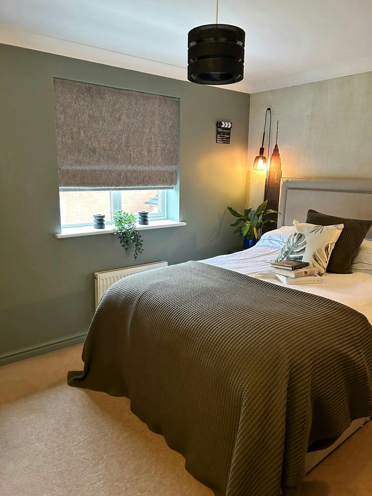 A bed covered with a green blanket is in a cozy bedroom. It rests beside a window with blinds and two potted plants. A hanging lamp adds warm light.