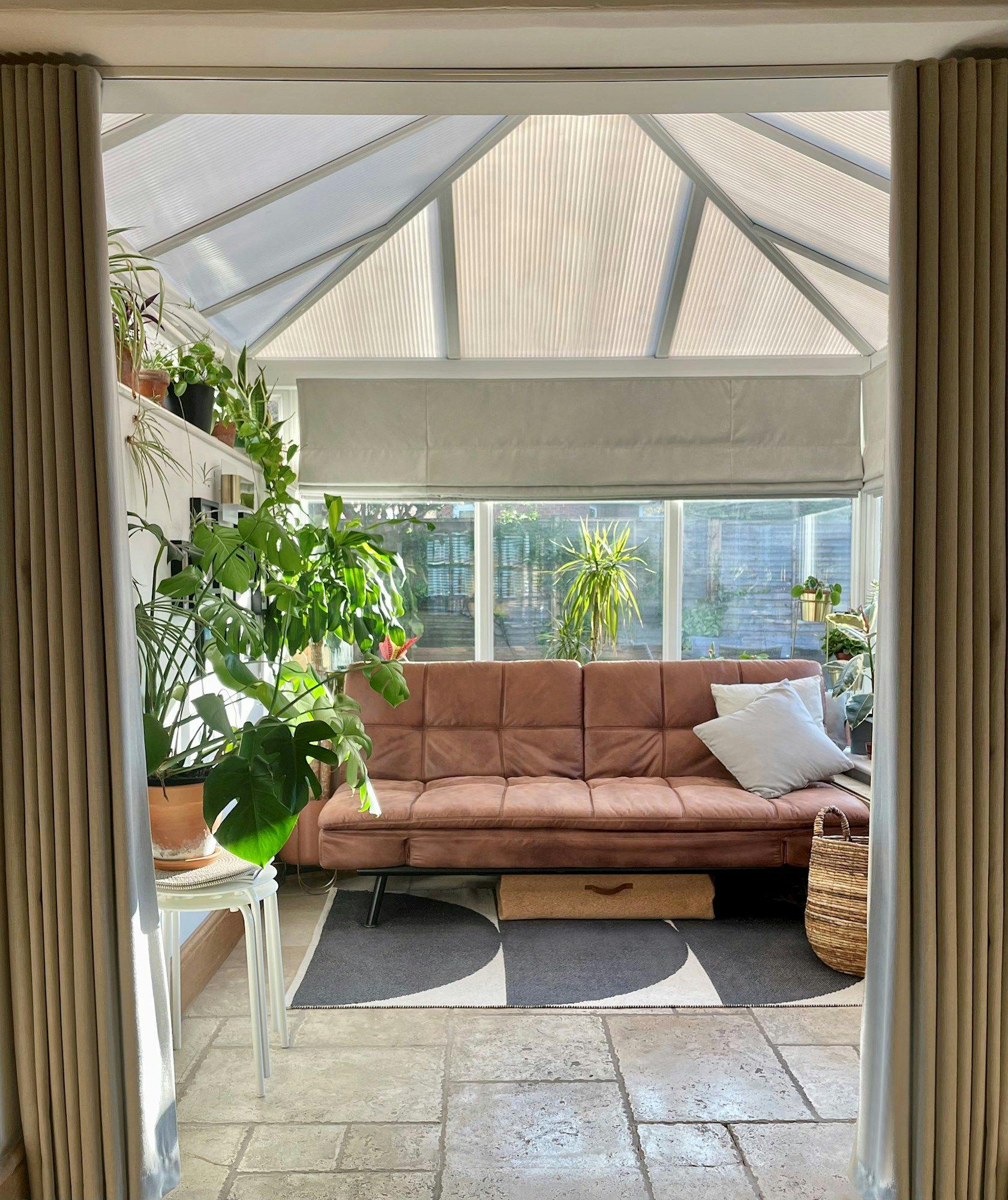 A brown sofa sits in a sunlit room with a glass roof. Surrounding it are various potted plants on shelves and tables, adding a lush, green atmosphere.