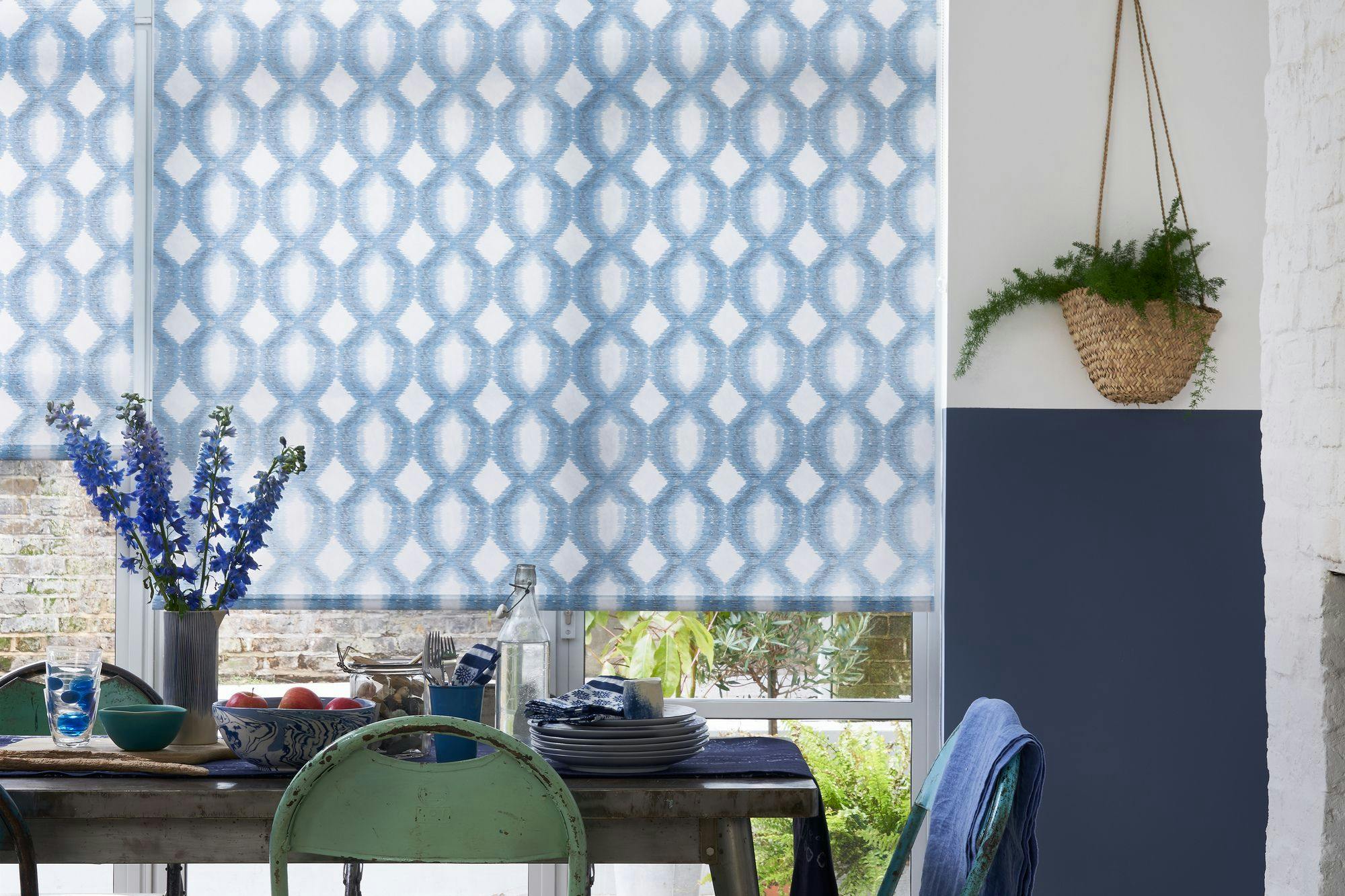 Window shades with a blue geometric pattern cover a window above a table set with plates, a vase with blue flowers, utensils, and decorative bowls. A hanging plant decorates the wall.