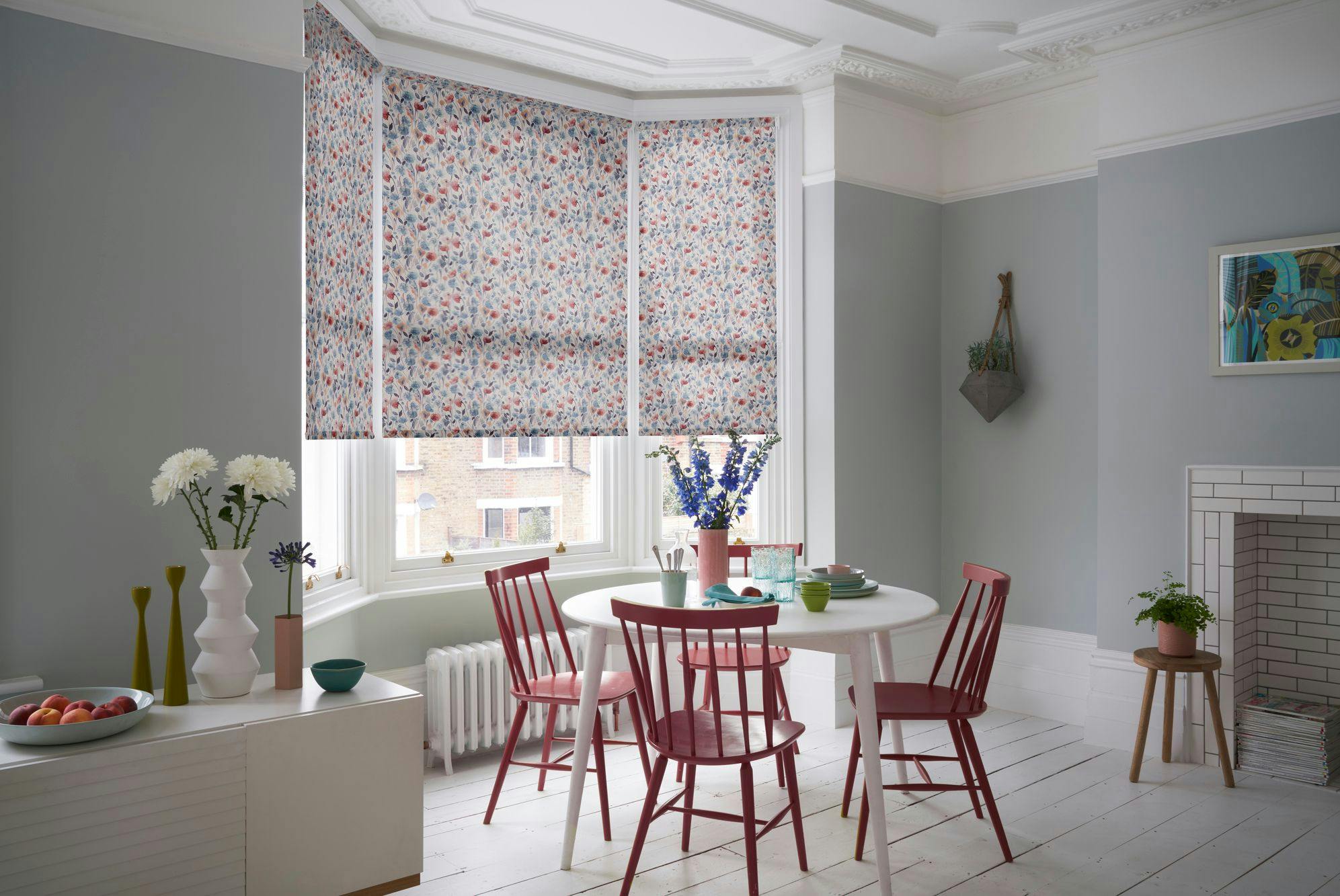 Dining table set with red chairs and tableware, surrounded by gray walls and floral-patterned window blinds. Vases with flowers adorn the area, enhancing the cozy, elegant room decor.