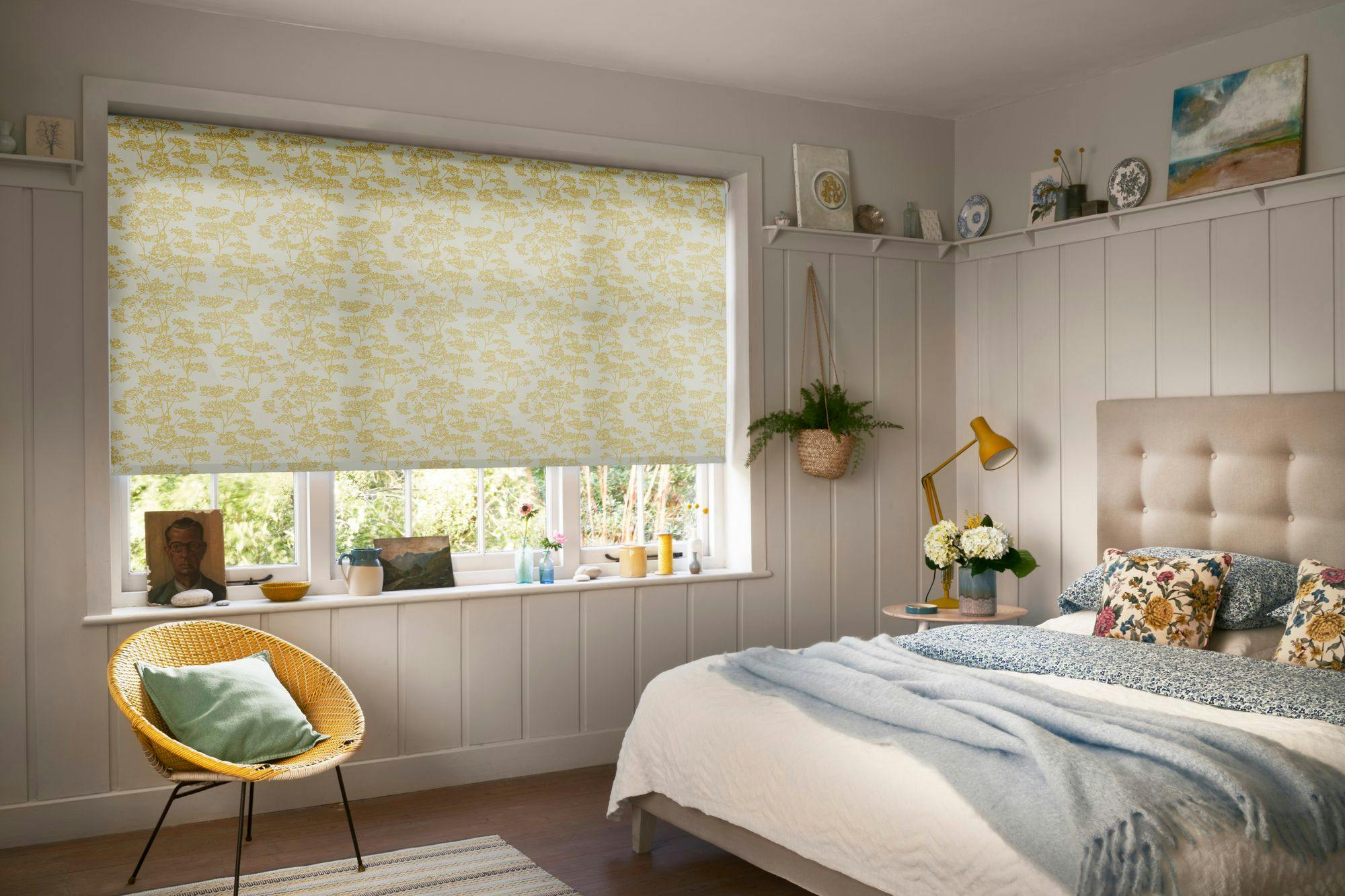 A cozy bedroom featuring a bed with floral pillows, a light blue blanket, and a yellow lamp on a wooden nightstand. A window has yellow-patterned blinds. A wicker chair holds a green cushion.