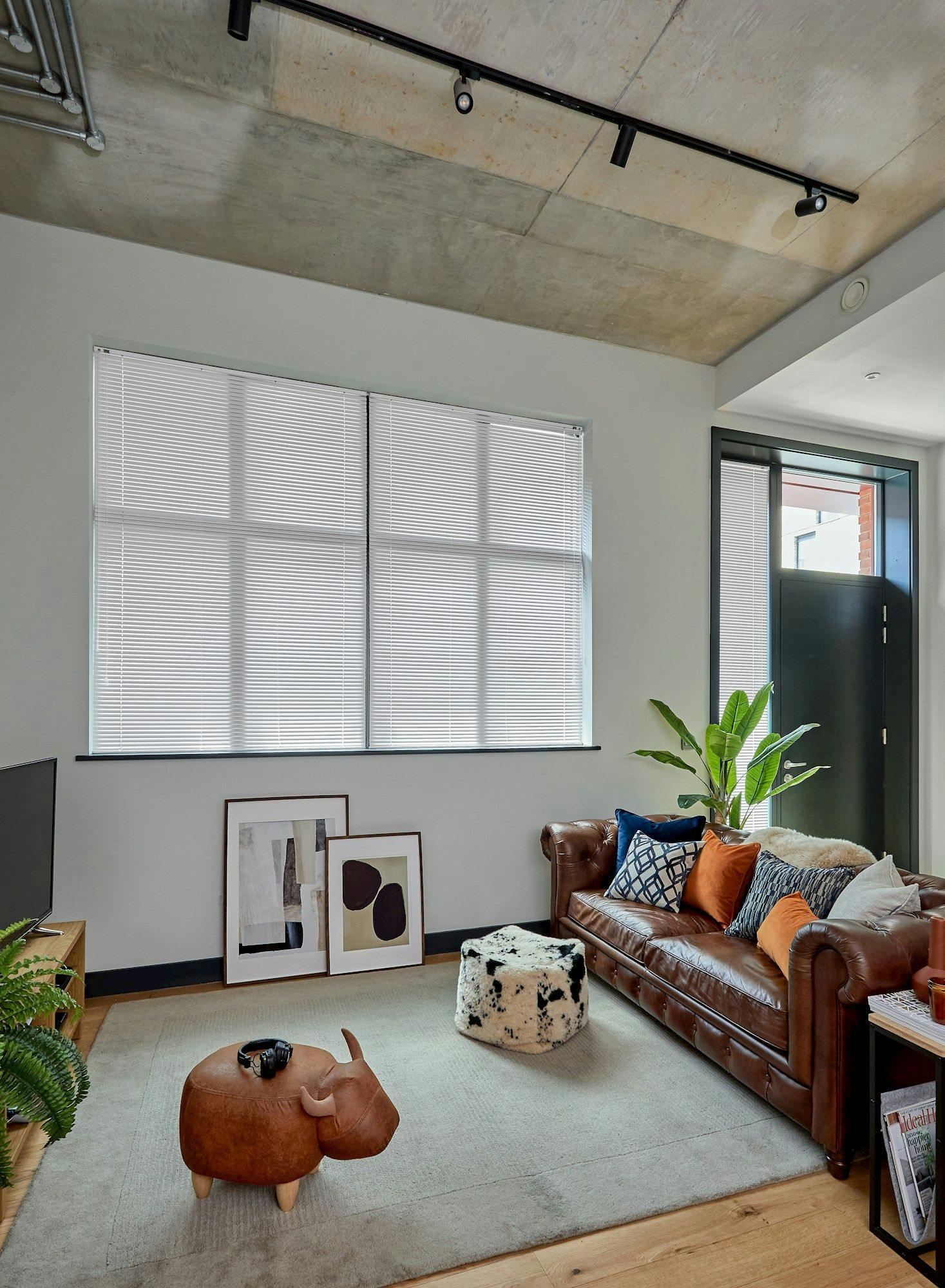 A brown leather sofa with colorful cushions sits in a modern living room. Nearby, a unique animal-shaped ottoman and abstract art pieces rest on the floor. Large windows provide natural light.