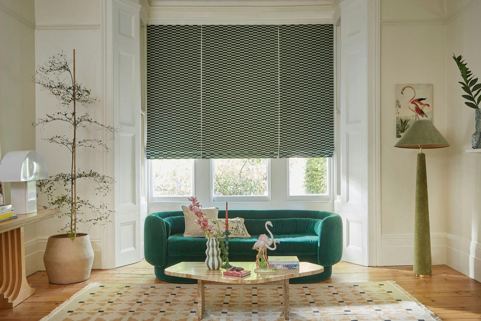 A green sofa sits against a window with patterned blinds, surrounded by a potted plant, a large green lamp, a coffee table with books and decor, and a framed flamingo picture.
