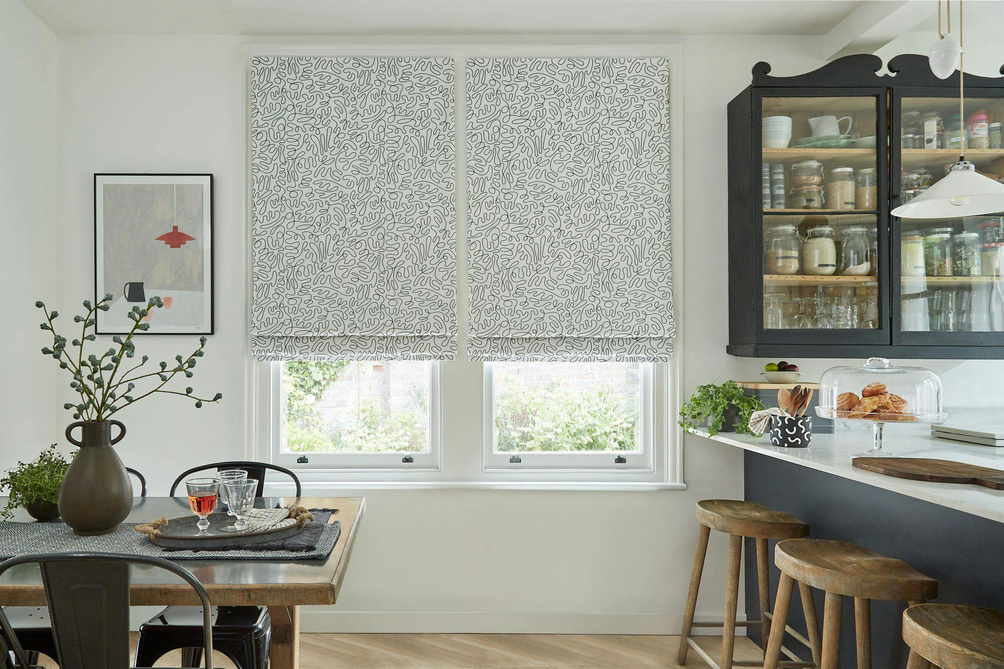 Table with plants and glassware rests in a cozy kitchen with patterned blinds covering windows. A black cabinet holds jars, and wooden stools line the counter. A framed picture hangs nearby.