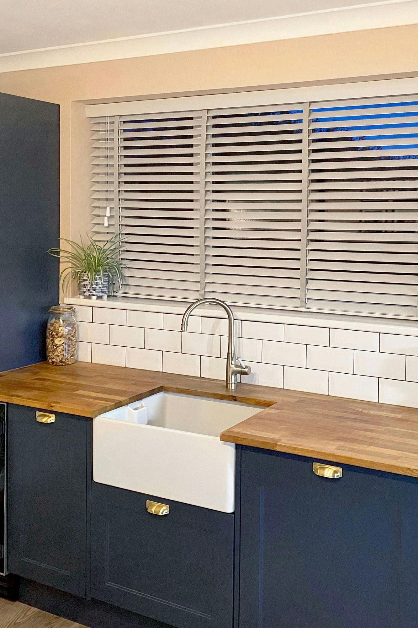 A kitchen sink with a chrome faucet sits in wooden countertops, under white tile backsplash. A jar and potted plant decorate the area; window blinds cover a large window above.