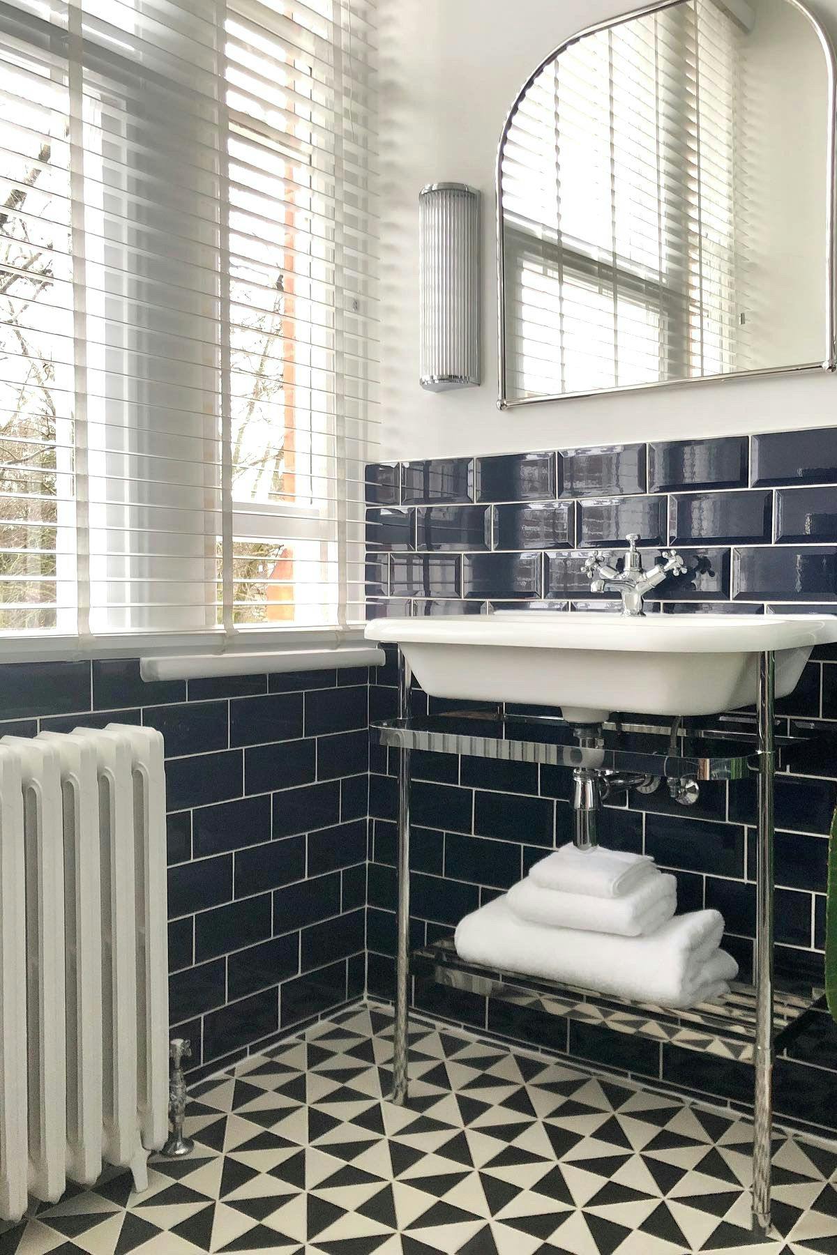 A white sink with a chrome faucet stands against a dark subway tile wall. It is beneath a large mirror, next to a window with blinds. Geometric-patterned black and white floor tiles enhance the space.