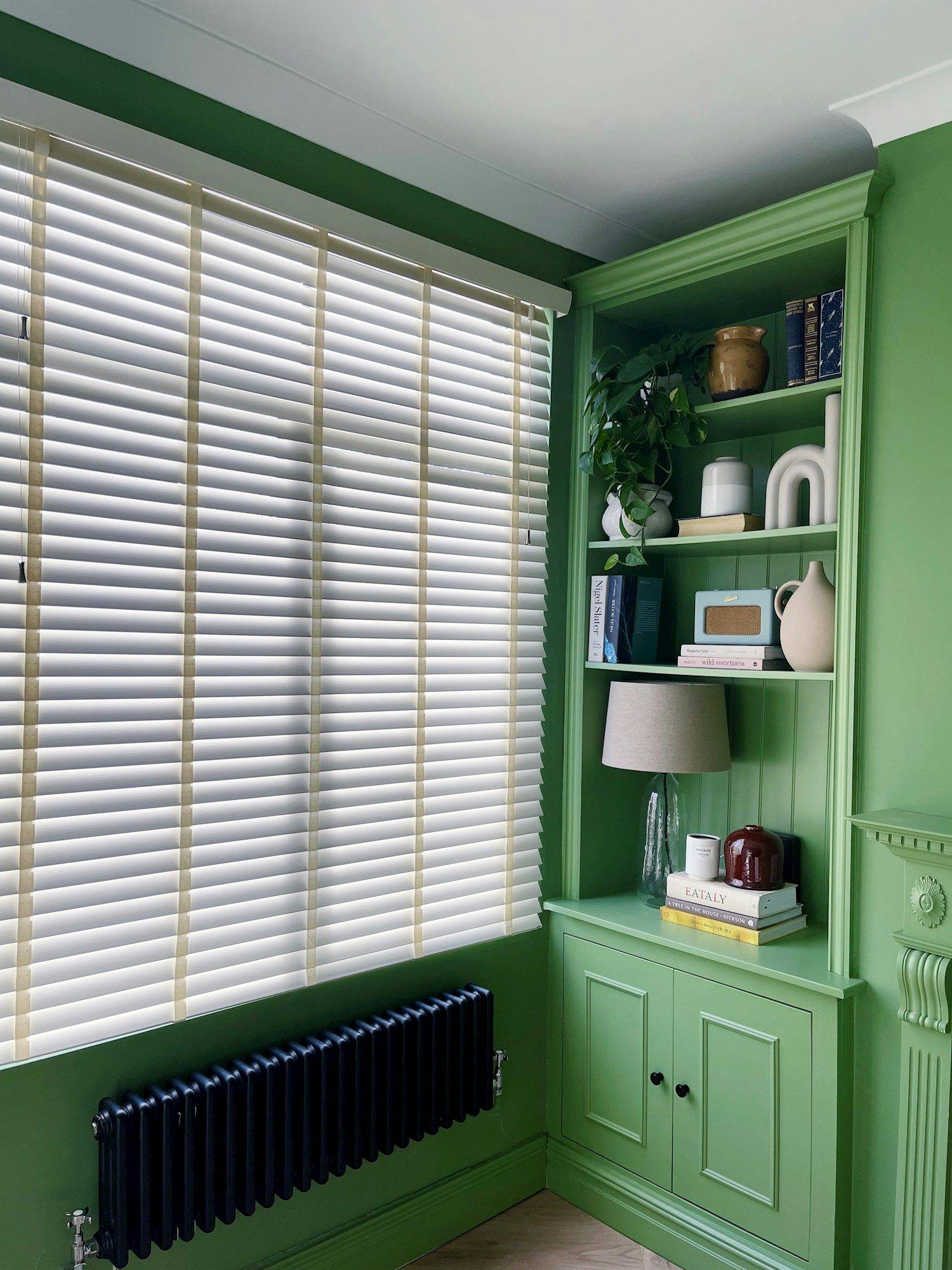 A green corner shelf holds books, a lamp, and decor items beside a window with white blinds. A black radiator is below, enhancing the vibrant room setting. Books read: "EATALY" and "MOR."