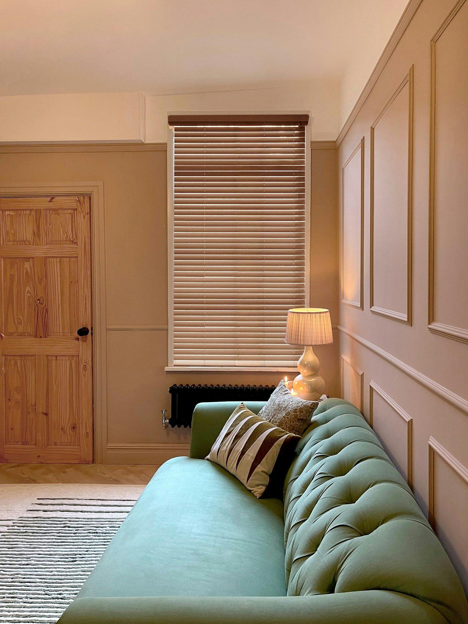 A green tufted sofa sits against a paneled wall, adorned with patterned cushions; a lit table lamp rests on a side table beside a window with closed blinds and a wooden door.