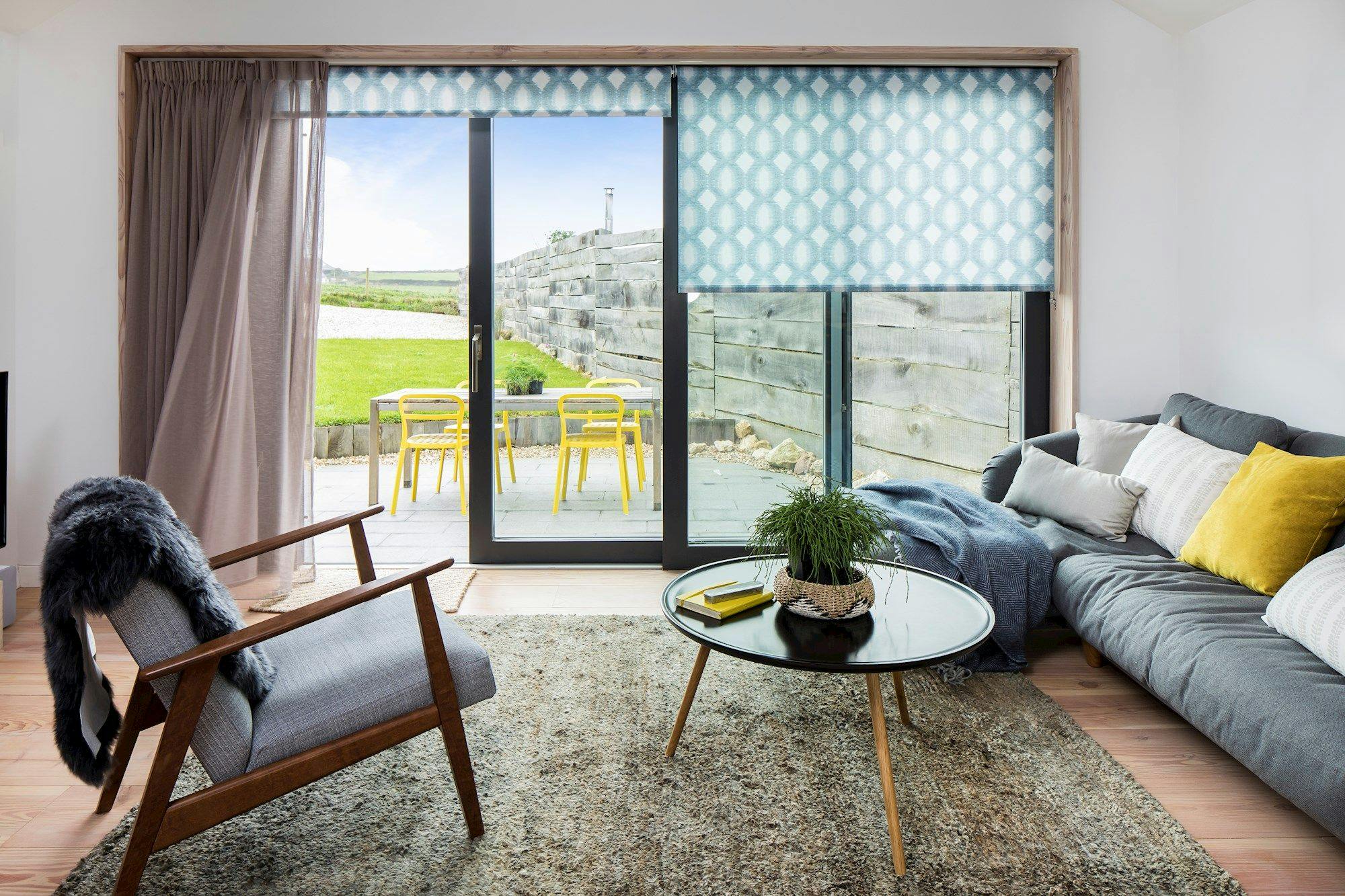 A modern living room features a gray sofa with colorful pillows and a round glass table with plants. Through sliding glass doors, a patio with yellow chairs and grassy yard is visible.