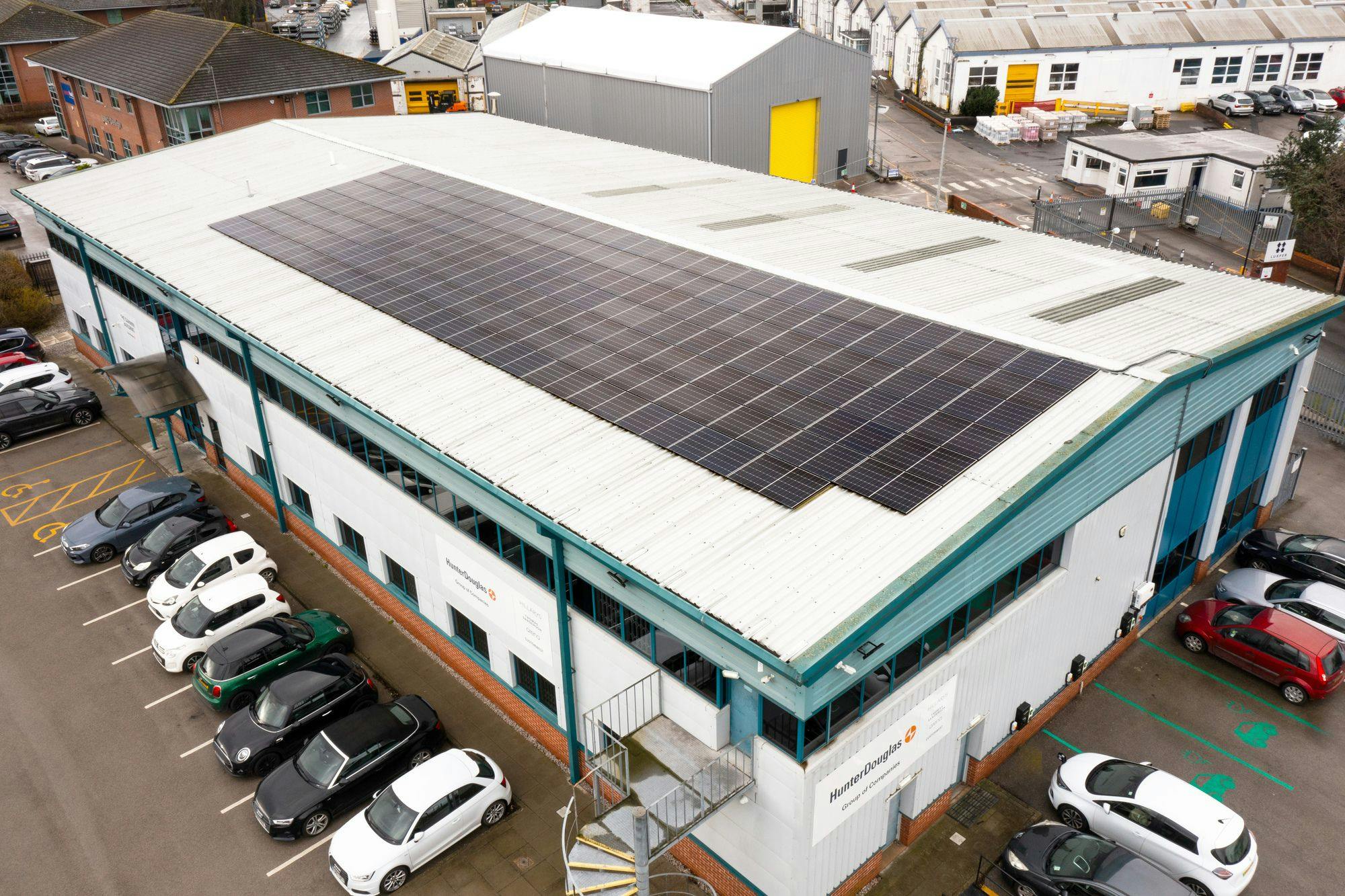 Solar panels cover a large industrial building roof surrounded by parked cars. Signs read "HunterDouglas." Nearby are other industrial buildings and parked cars.