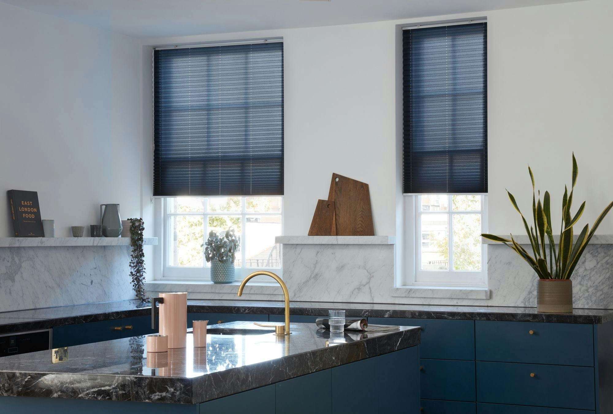 Kitchen features a marble countertop with pink canisters and a gold faucet. Two windows have black blinds. Items include a book titled �EAST LONDON FOOD,� cutting boards, and plants.