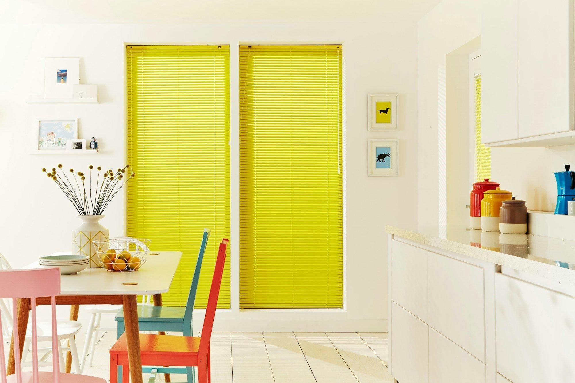 Table decorated with colorful chairs, a vase of flowers, and a fruit bowl sits near windows with bright yellow blinds in a cozy, modern dining room. Walls feature small framed art.