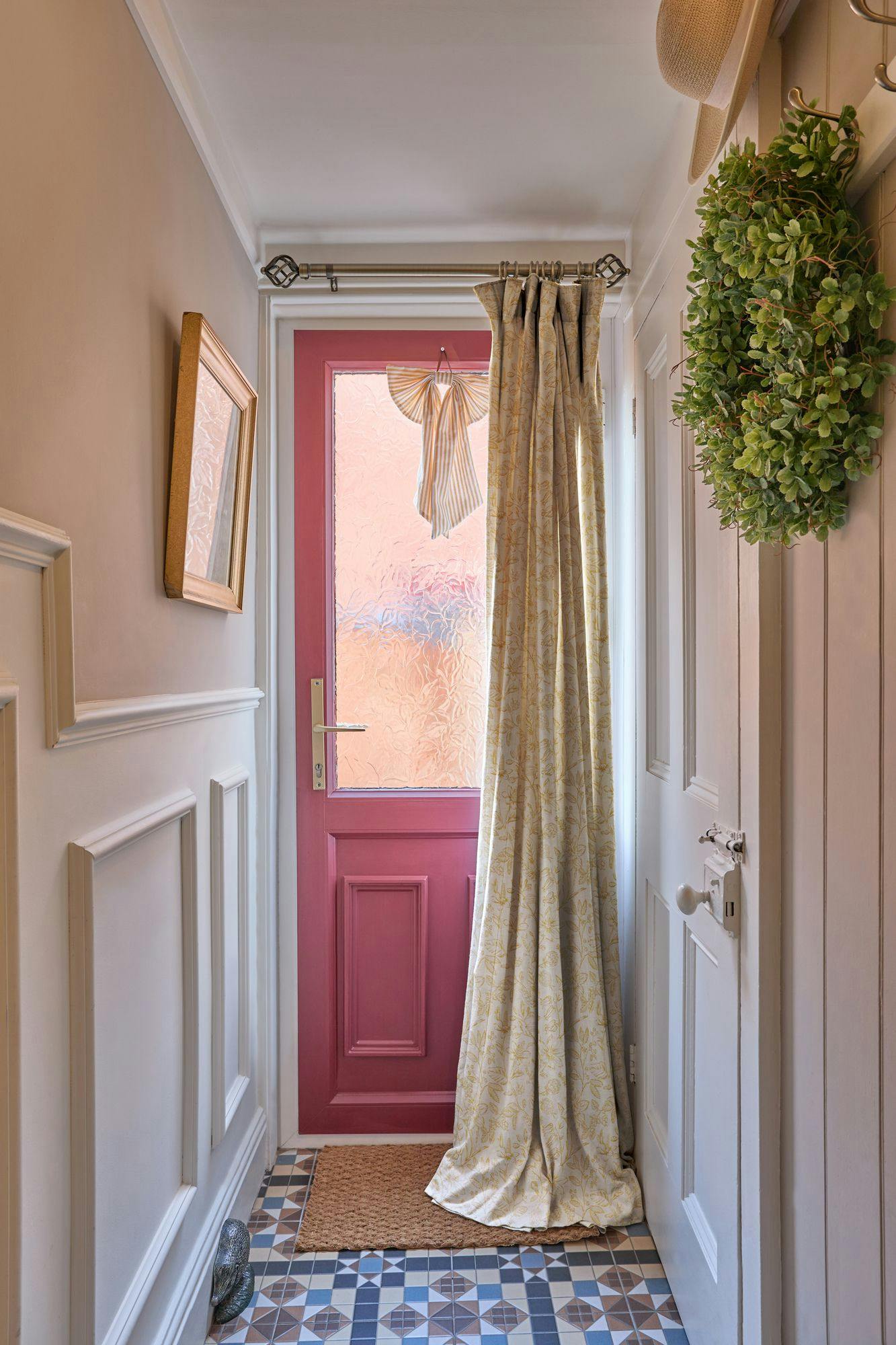 Pink door adorned with frosted glass, partially covered by patterned curtain in narrow hallway. Tiled floor beneath. Green wreath and straw hat hang on opposite wall.