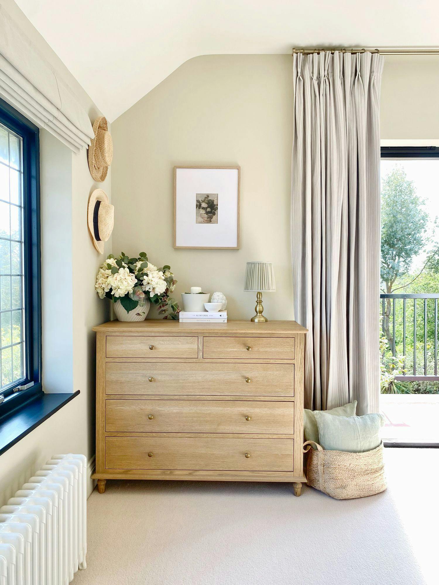 A wooden dresser stands against a cream wall, adorned with framed art, baskets, and flowers. A window with closed curtains and hanging hats completes the bright, airy room decor.