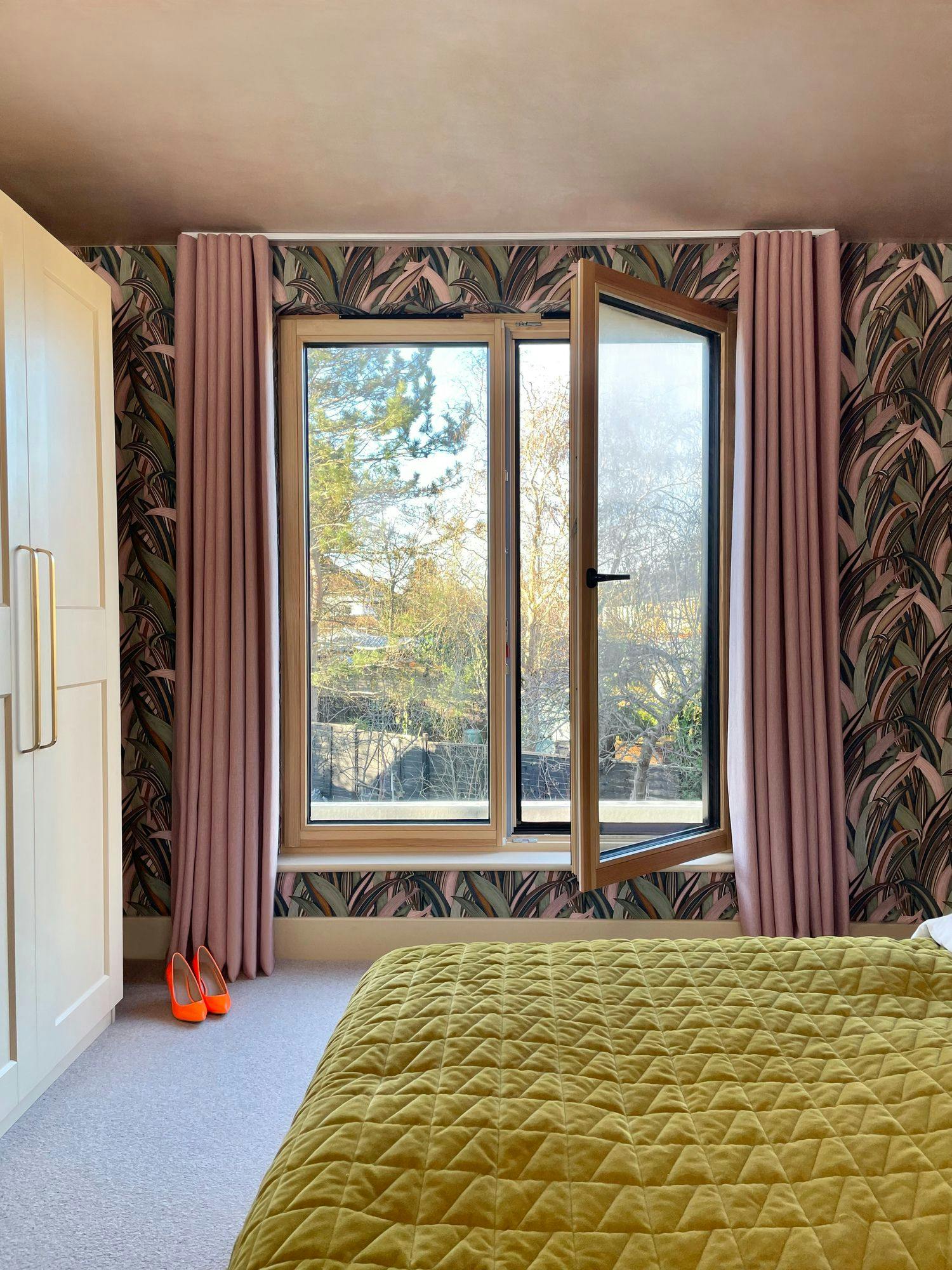 Open wooden-framed window lets light into a bedroom with pink curtains and patterned wallpaper. A mustard yellow quilt covers the bed, and orange high heels rest on the floor.