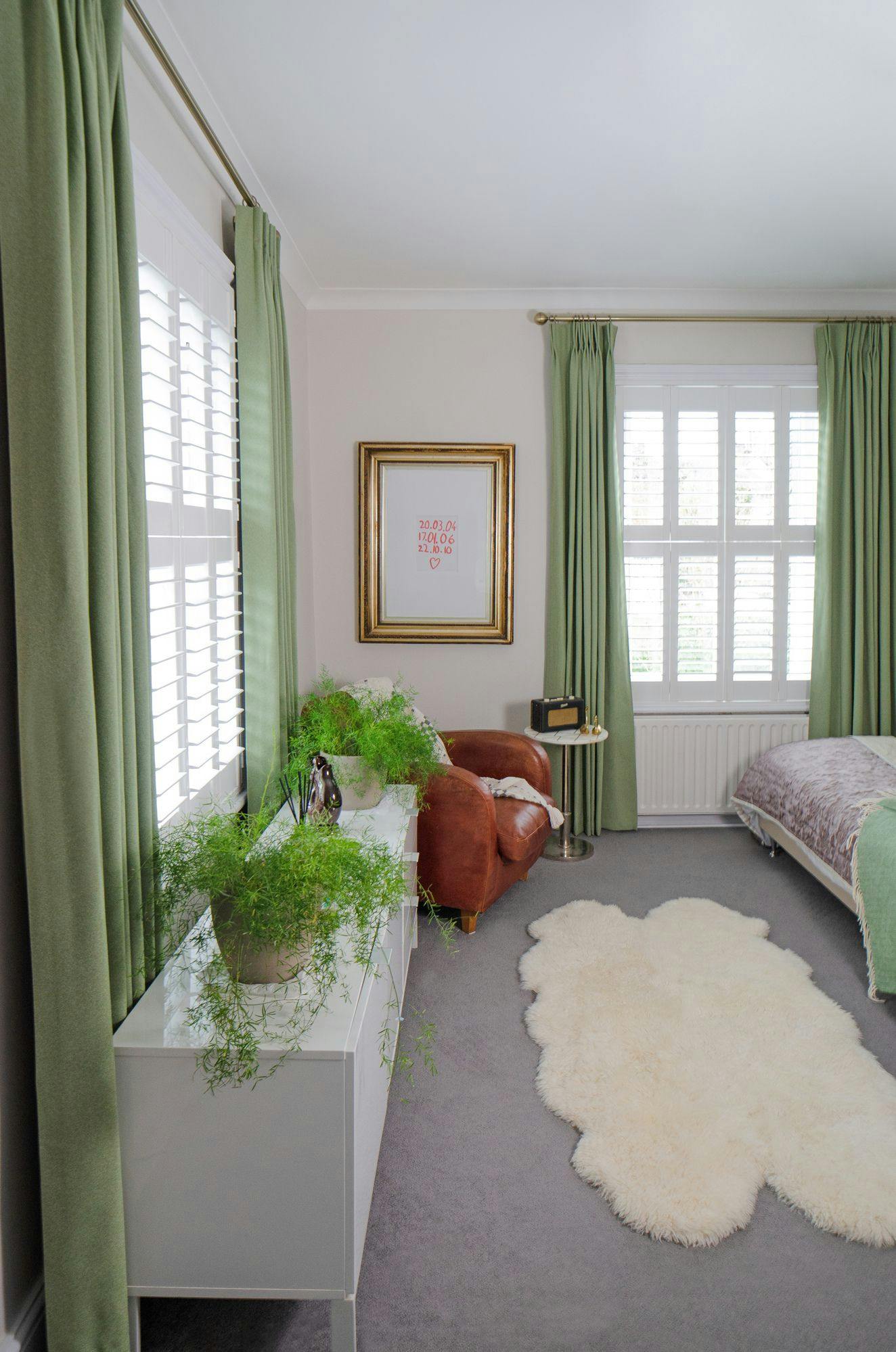 A cozy room features a brown leather chair beside a framed picture with red text "20.03.04, 17.01.06, 22.10.10 ??". A white cabinet holds lush plants. Green curtains frame the windows.