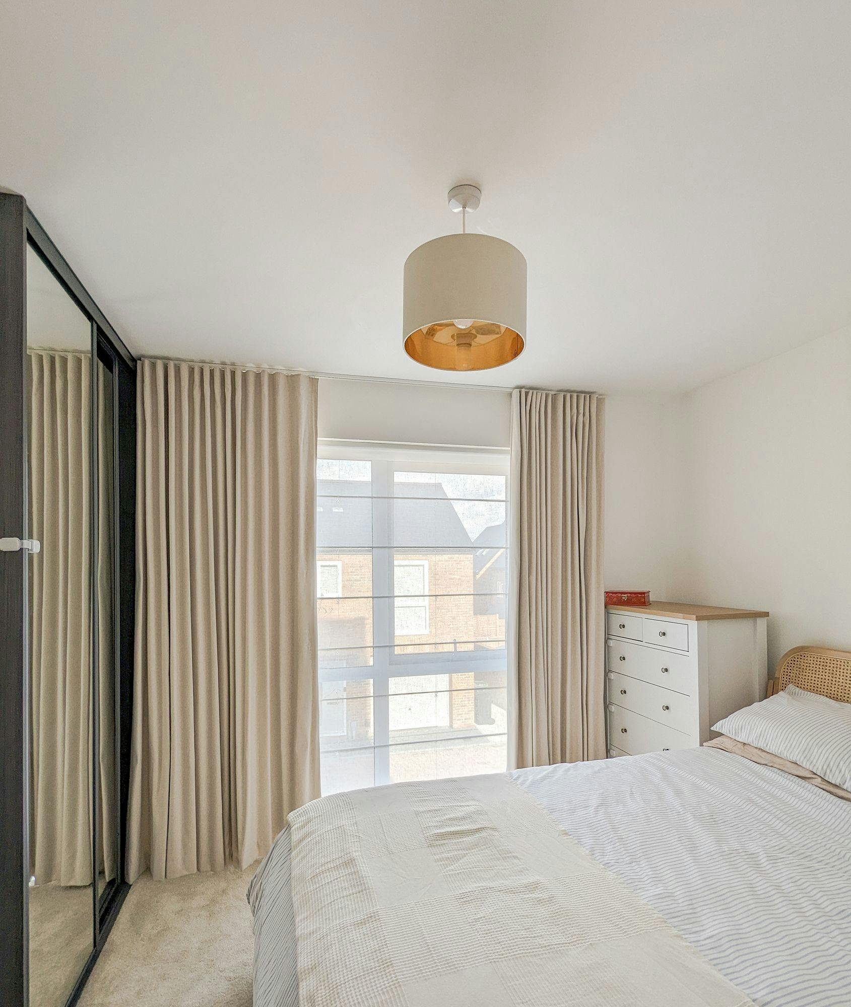 Bed positioned in a bright room with beige curtains, mirrored wardrobe on the left, white dresser on the right, and a simple ceiling light fixture above. Large window shows a view outside.