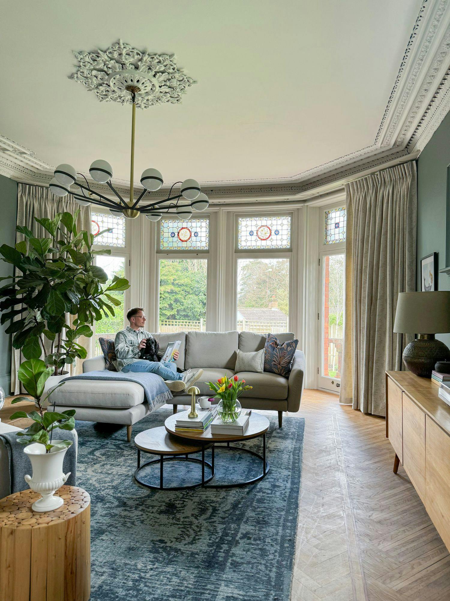 Man sitting on a gray sectional sofa, holding a dog, in a well-furnished living room. Stained glass windows and greenery enhance the bright space, with a chandelier overhead and coffee table nearby.
