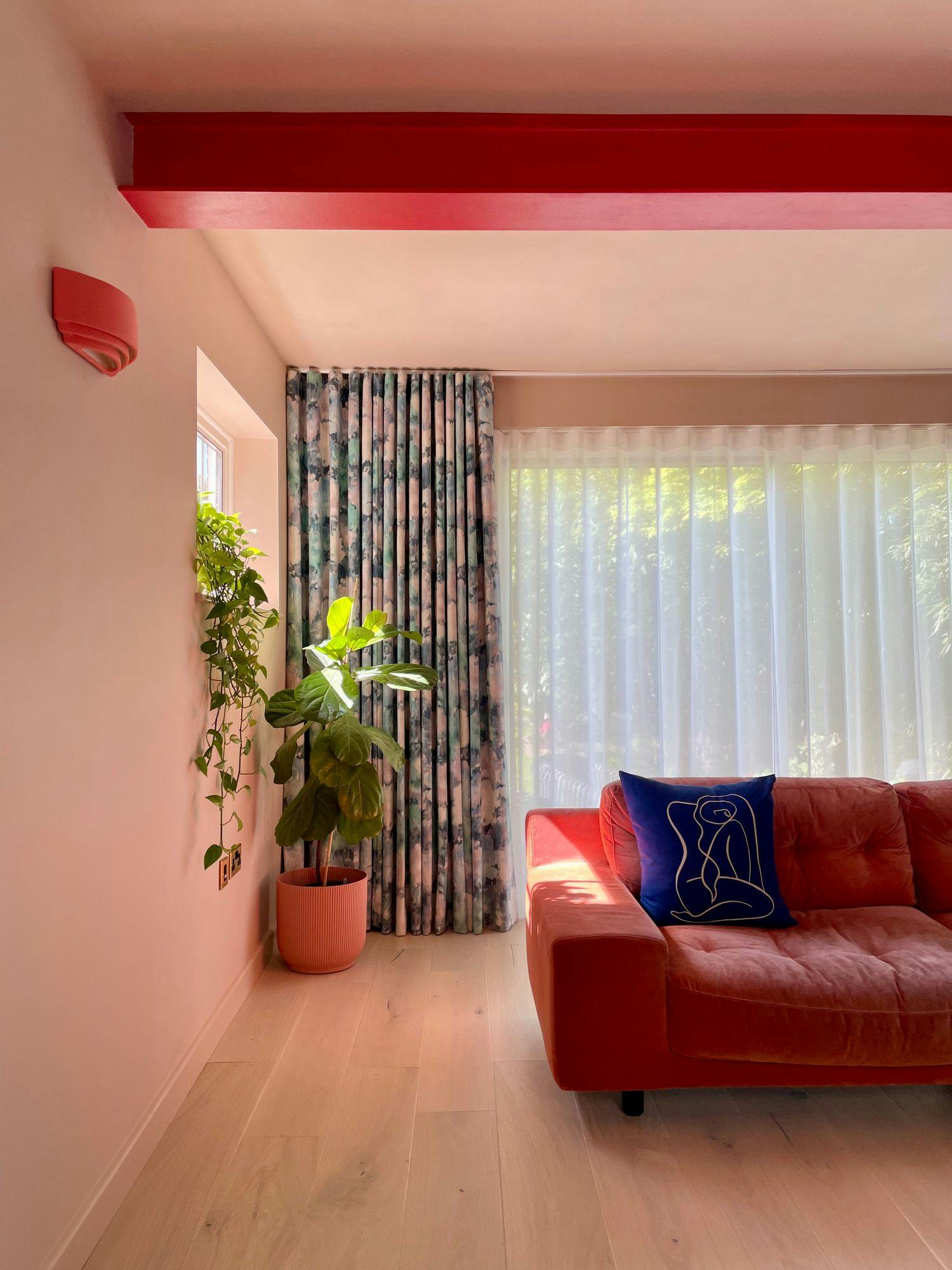 A red sofa holding a blue pillow sits in a brightly lit room with sheer curtains and floral drapes. Nearby, a green plant rests in a pink pot, under a red ceiling beam.