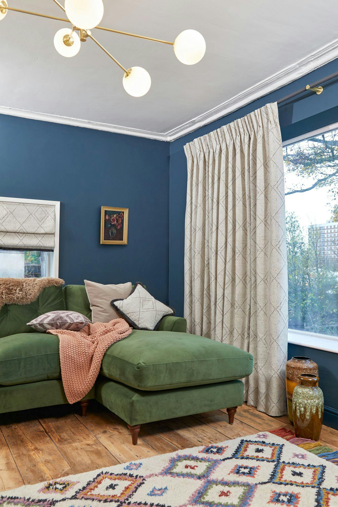 Green sectional sofa holds pillows and a knitted blanket. It faces a large curtained window. A wooden floor and patterned rug complement the space, illuminated by a modern chandelier.