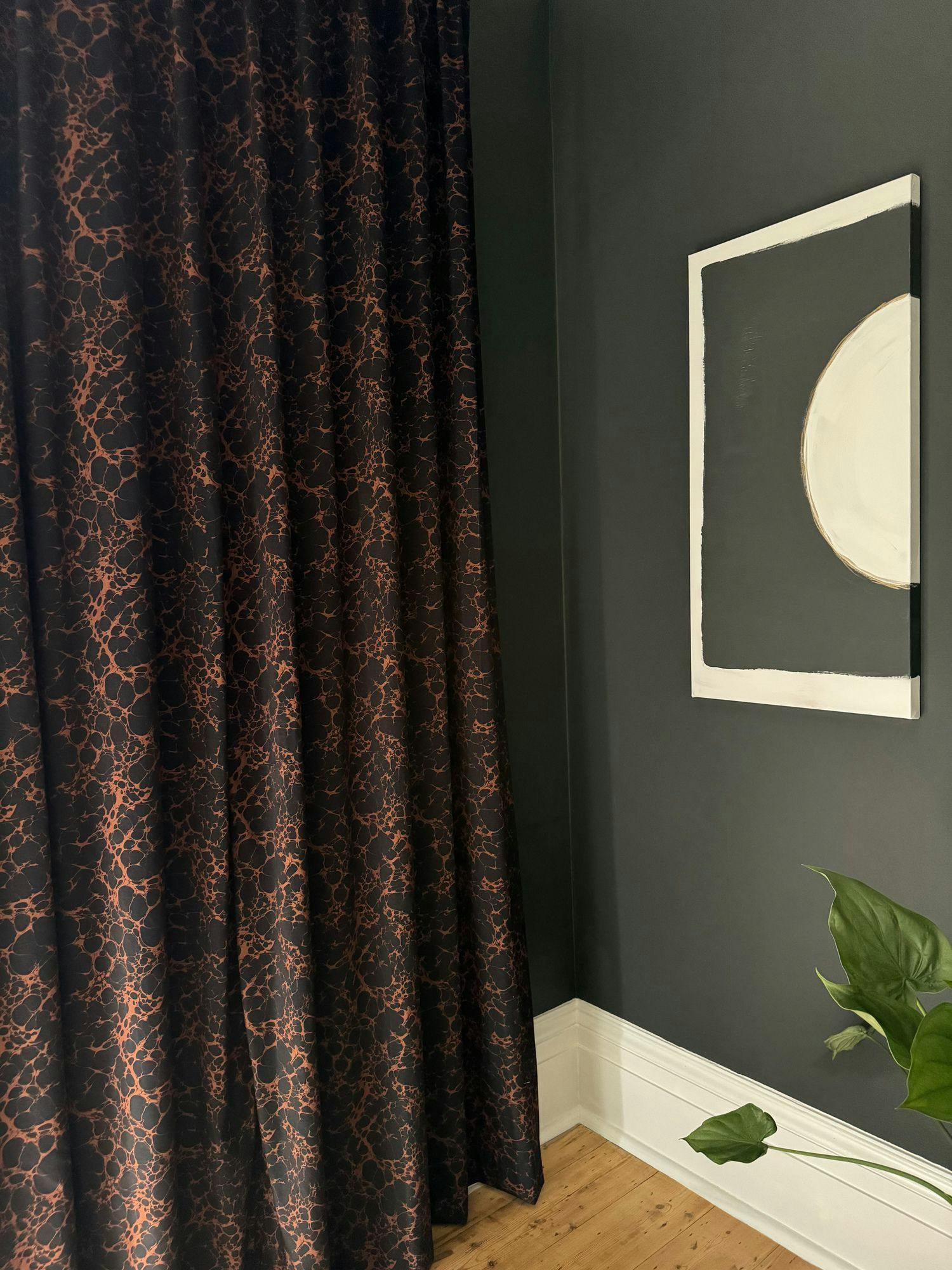 Patterned dark curtains hang beside a black wall with abstract circular artwork; a green plant sits on a wooden floor.