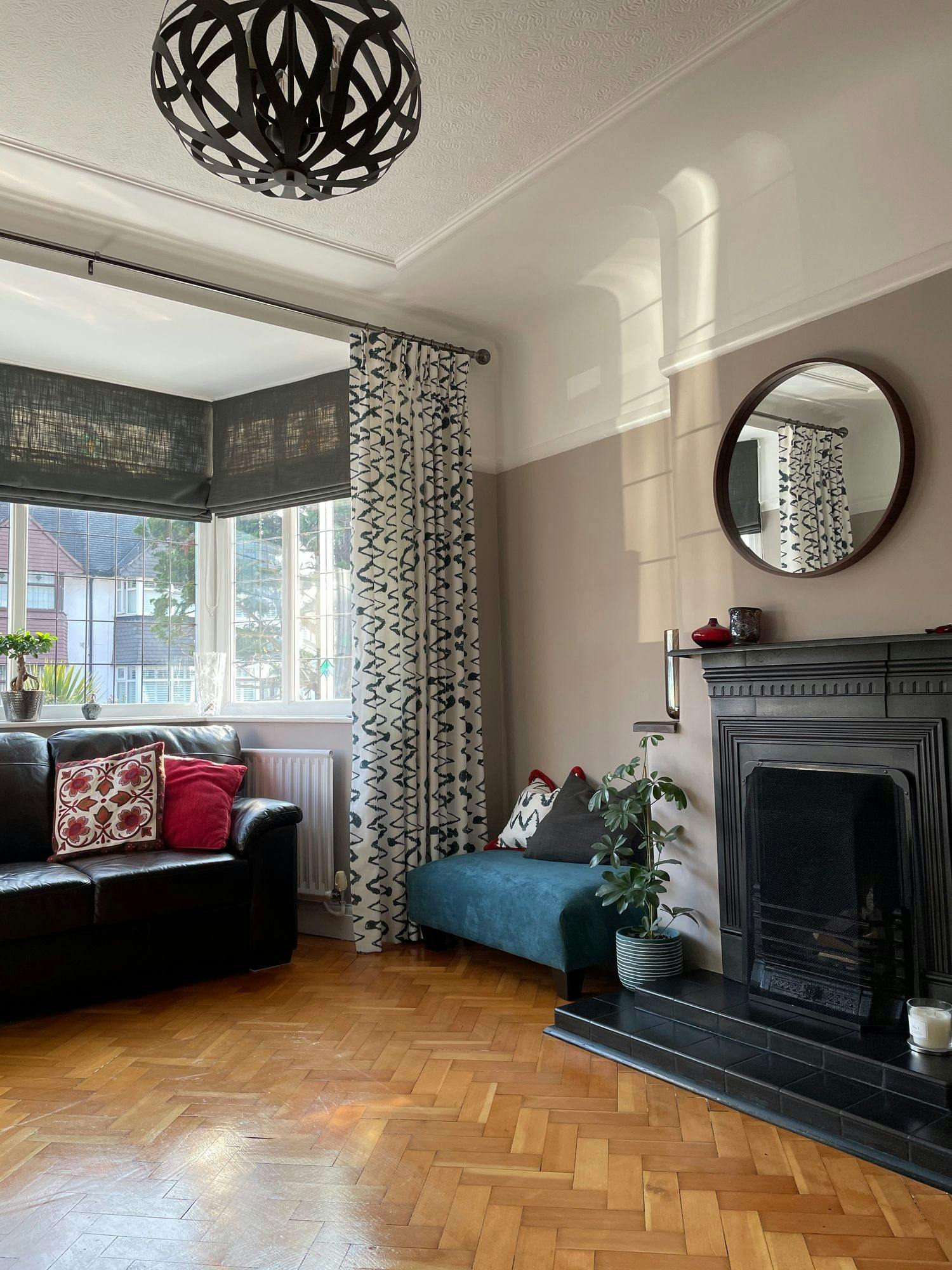 A living room features a black leather sofa with patterned cushions beside a large window with curtains. A potted plant sits by a fireplace, and a round mirror hangs above it.