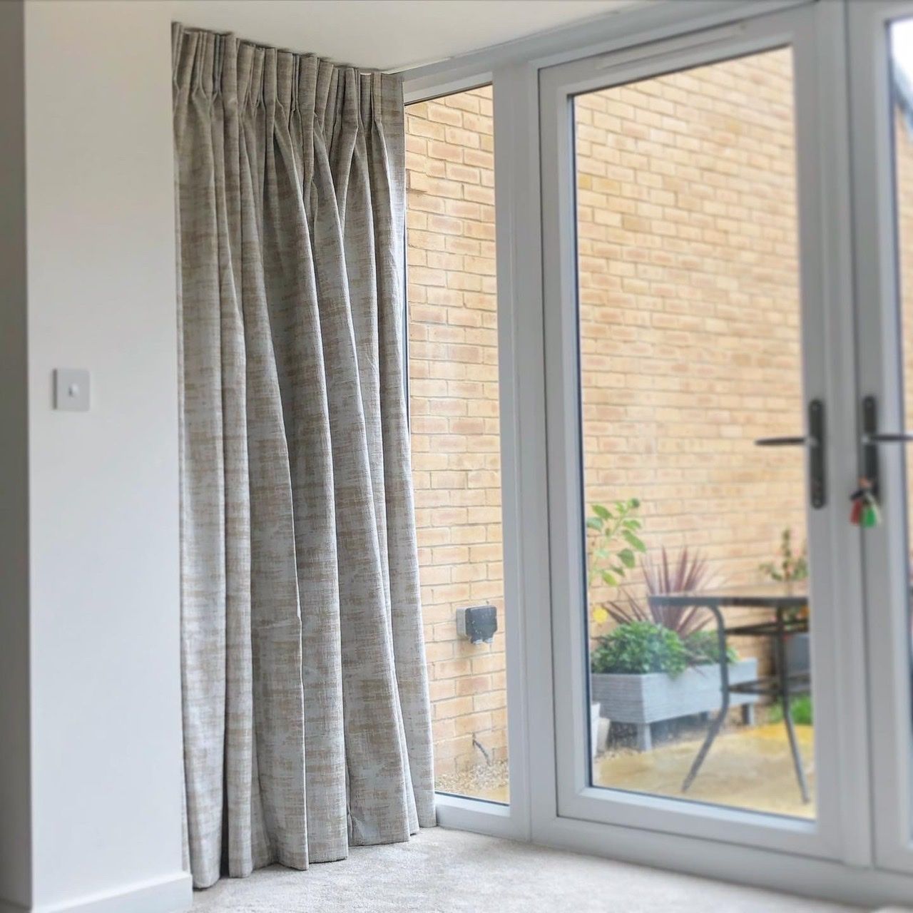 Curtains hang partially open beside a glass door, revealing an outdoor patio with a potted plant and brick wall in a bright, contemporary space.