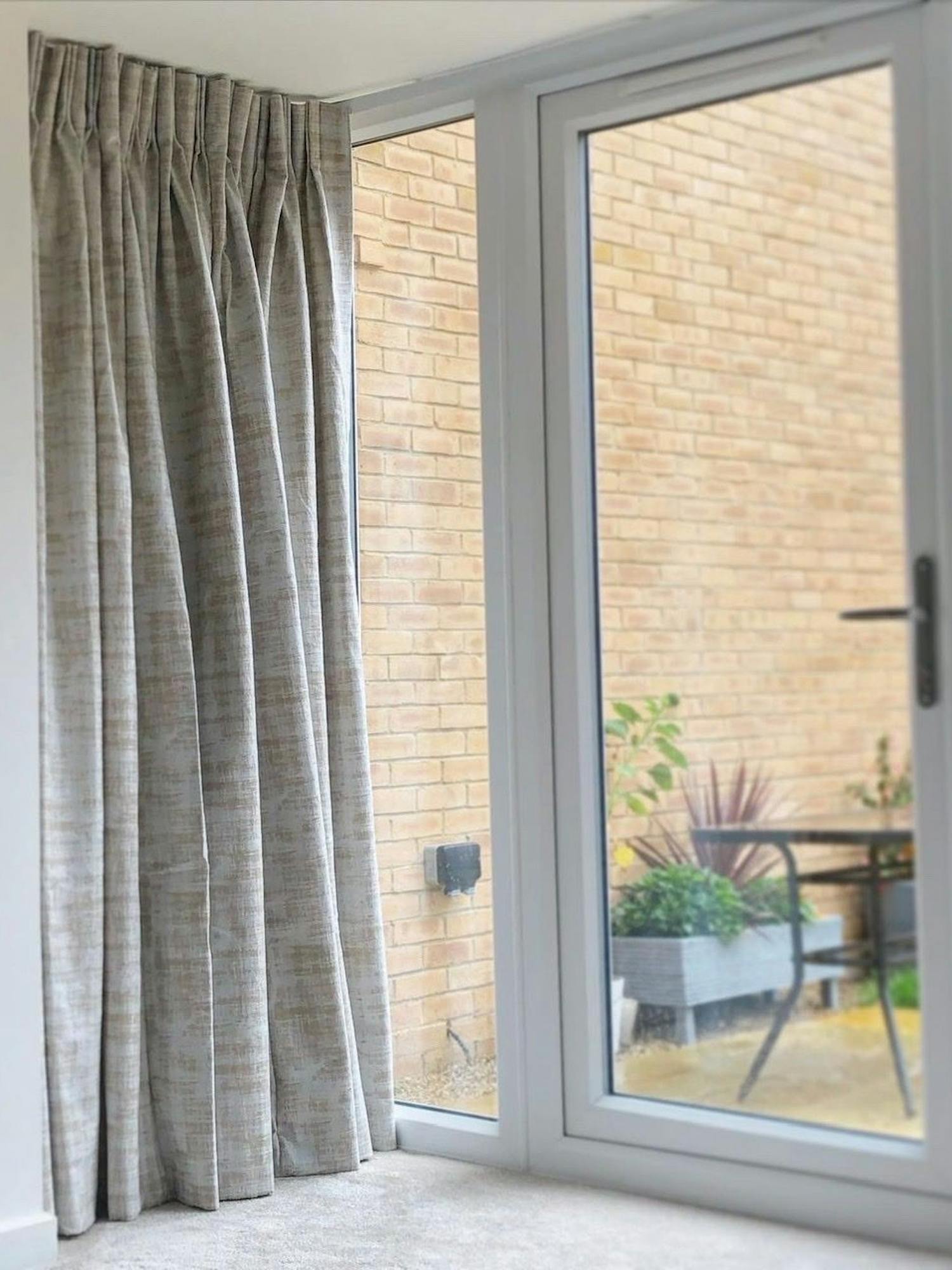 Curtains hang partially open beside a glass door, revealing an outdoor patio with a potted plant and brick wall in a bright, contemporary space.