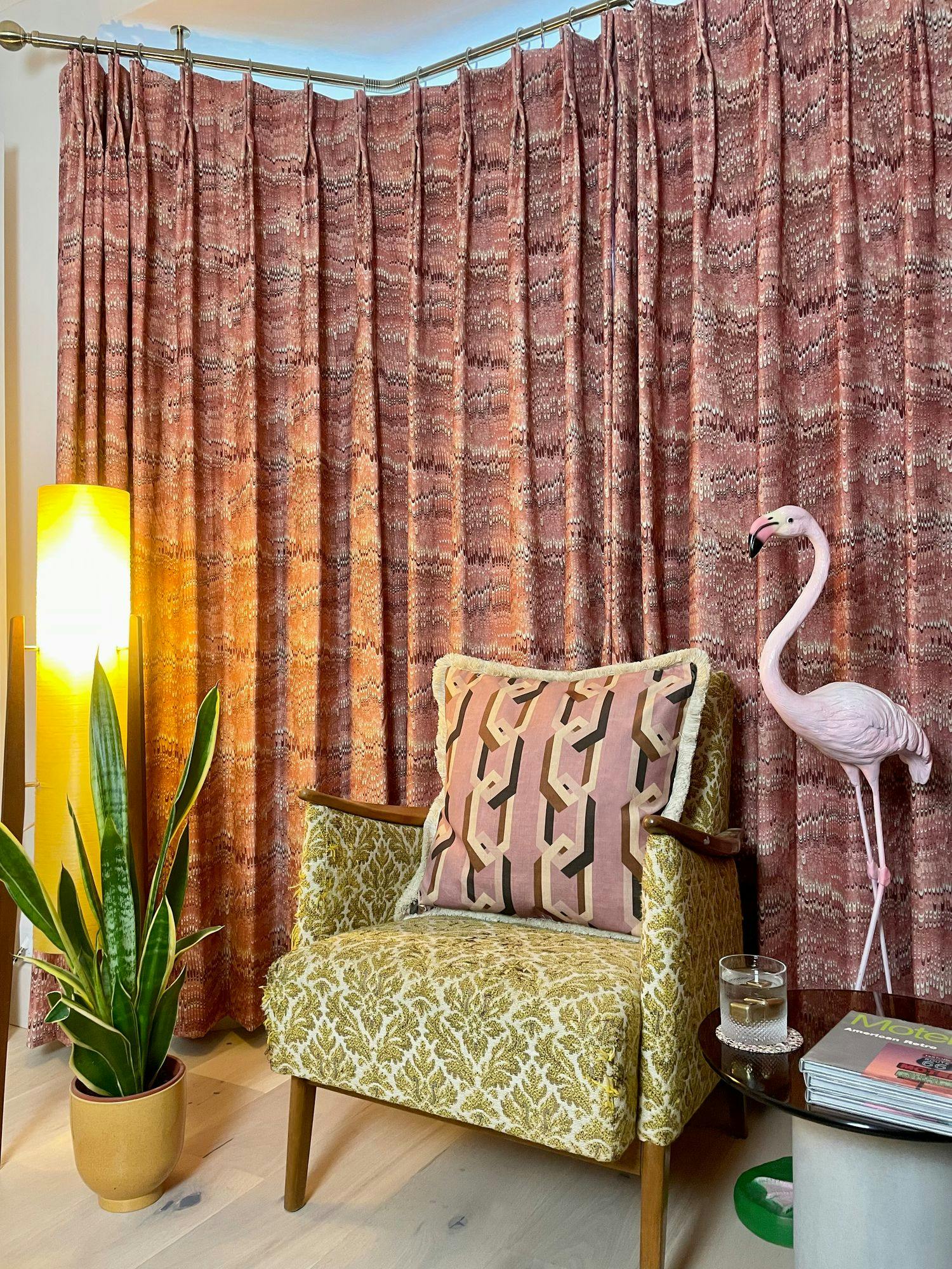 A patterned armchair with a geometric pillow sits beside a potted plant and illuminated lamp, against red-patterned curtains. A pink flamingo statue stands nearby atop a glass-top table with magazines.