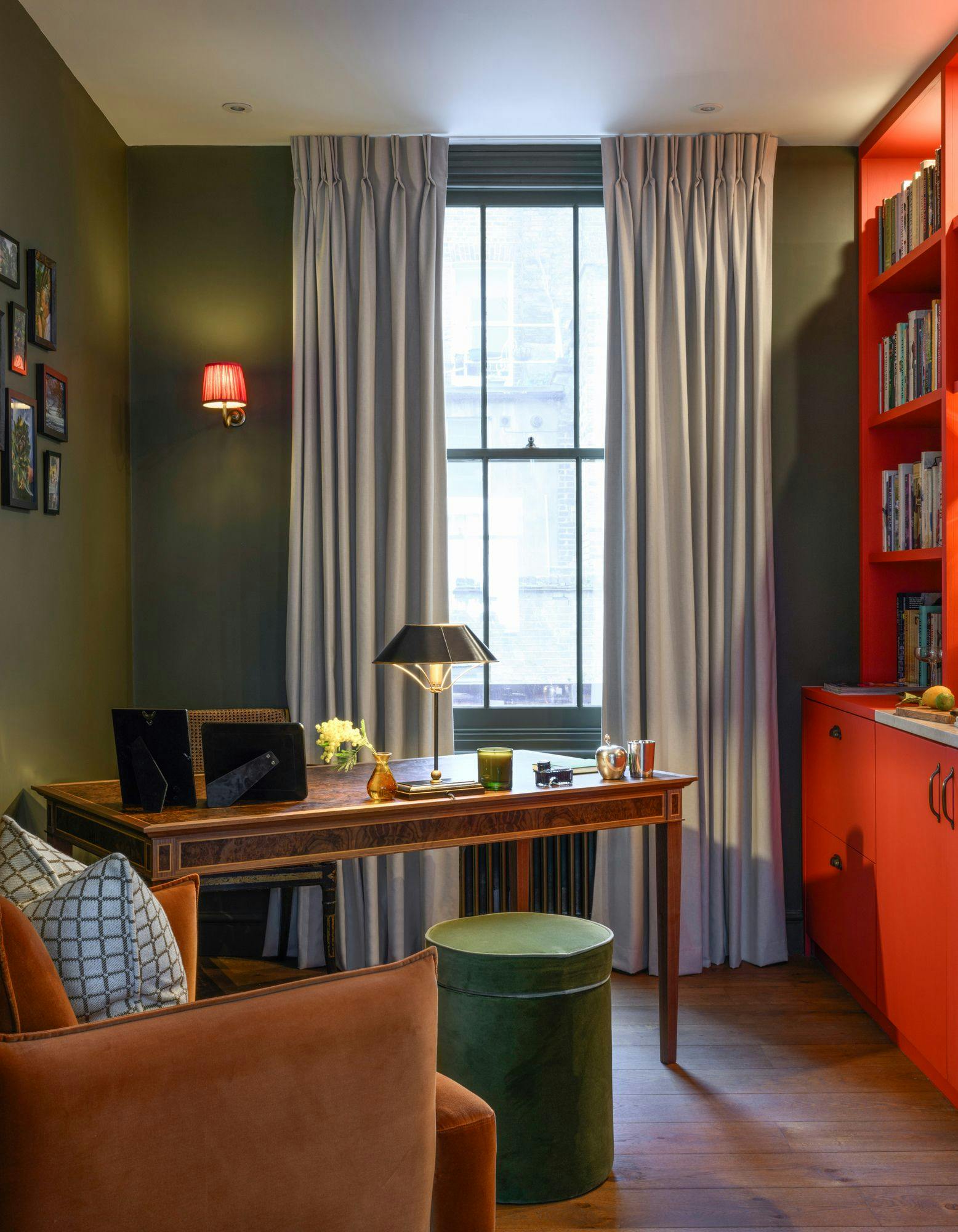 A desk holds a lamp, flowers, and framed pictures, situated in front of a curtained window. The room has olive walls, a red lamp, and a bright orange bookshelf filled with books.