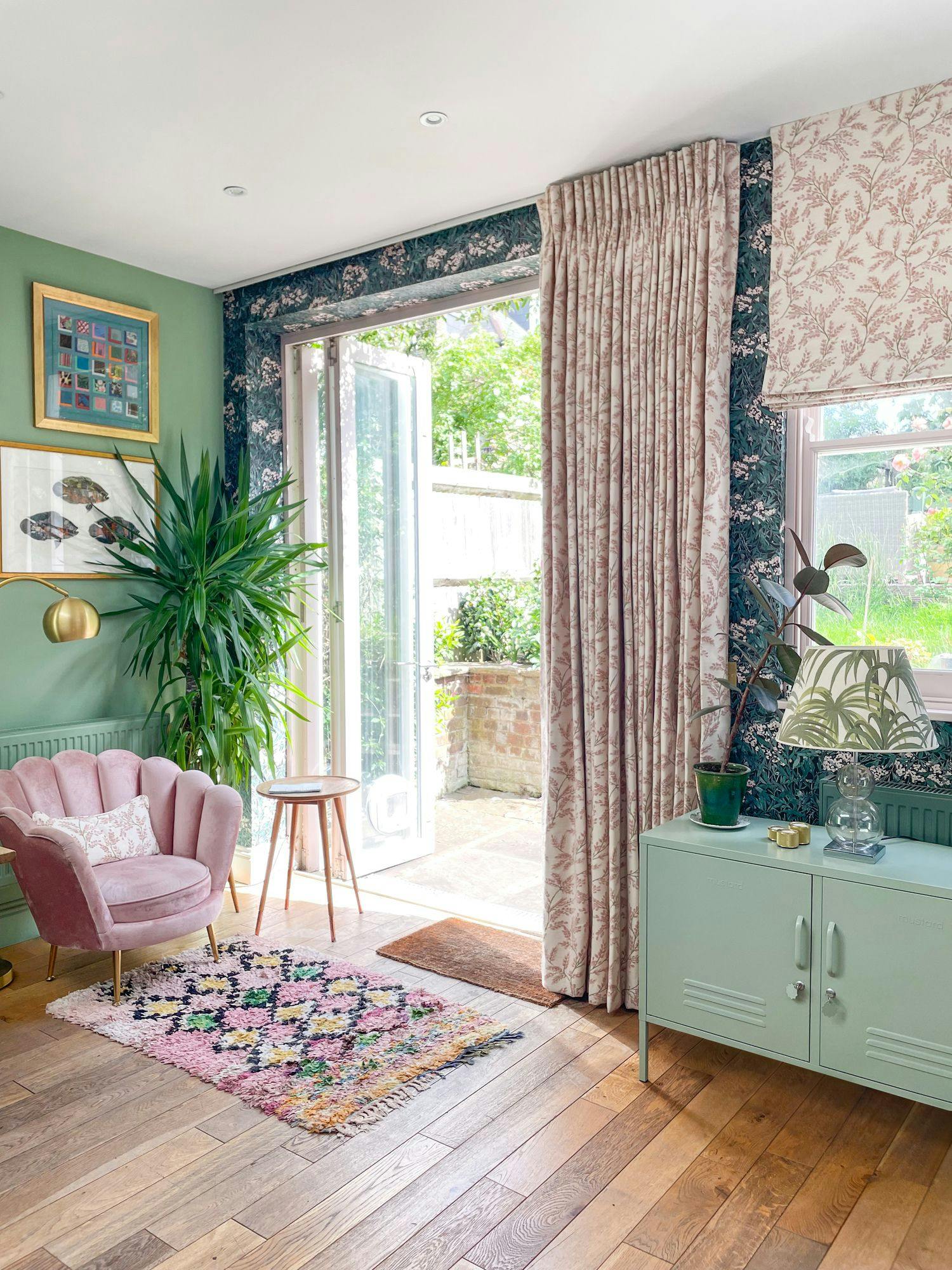 A pink armchair sits in a cozy, colorful living room with floral wallpaper and plants, facing open French doors leading to a garden. A green cabinet and vibrant rug accentuate the wooden floor.
