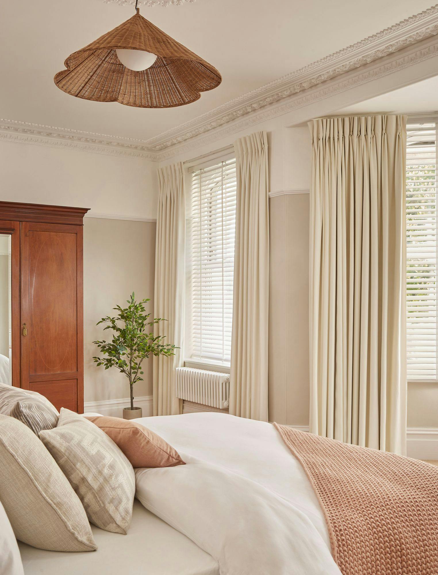 A neatly made bed with multiple pillows stands in a bedroom. A potted plant and a wooden wardrobe complement the cream curtains and blinds. A woven lampshade hangs from the ceiling.