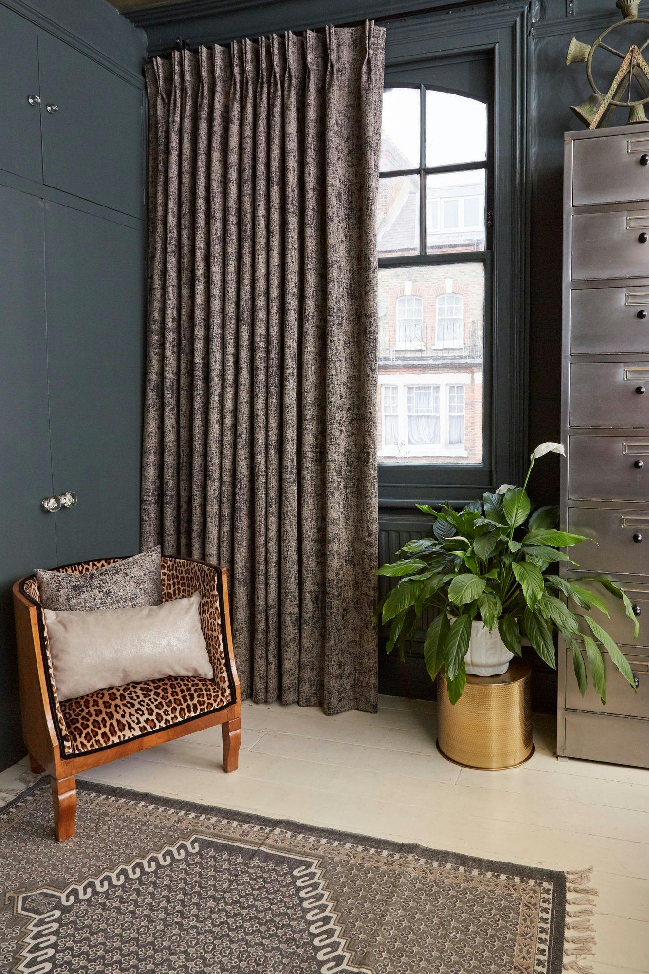 Chair with leopard-print upholstery and beige pillows sits near a potted green plant and a metal cabinet. Heavy curtains cover a large window, with intricate patterned rug on the floor.