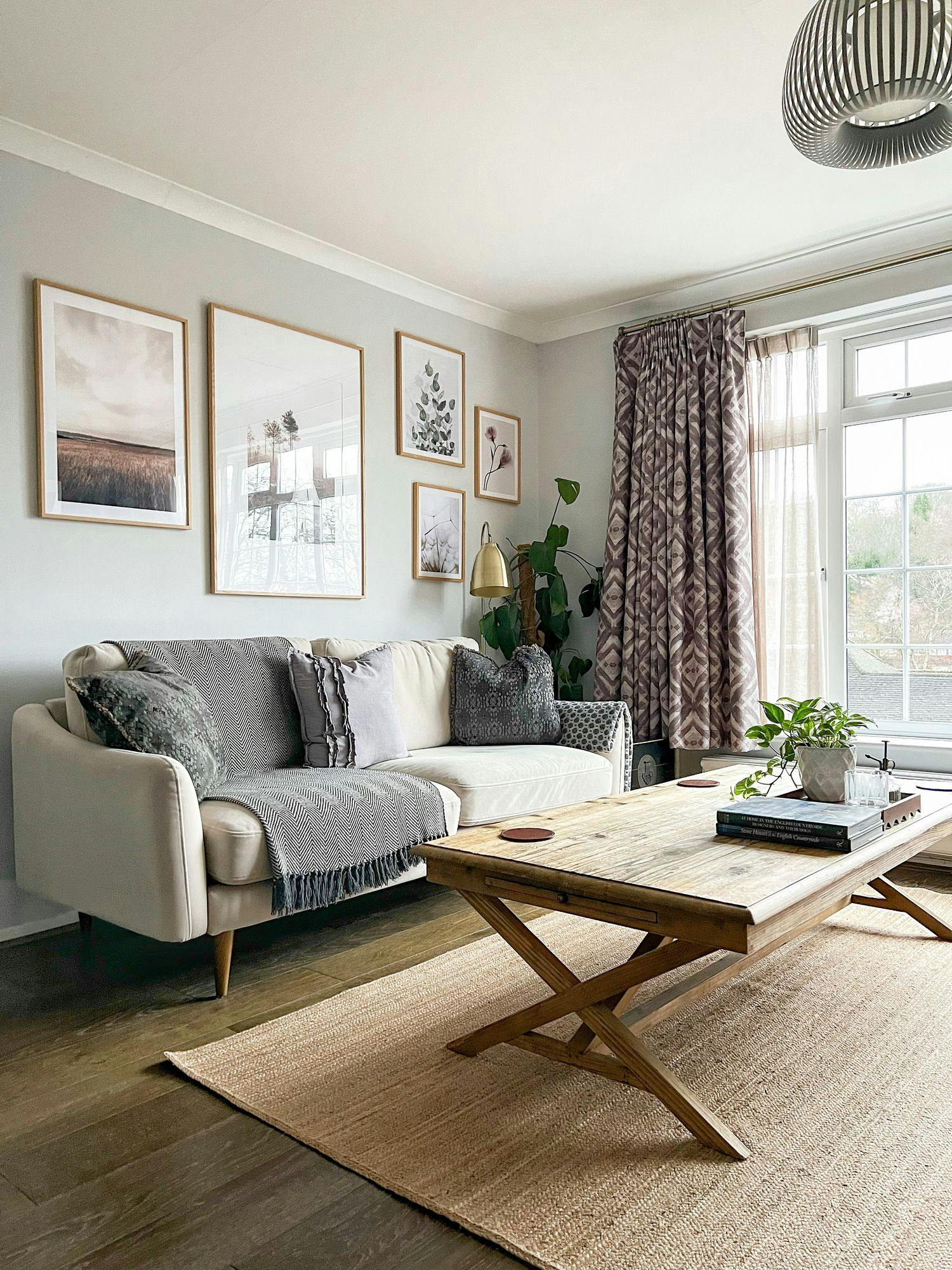 A beige sofa adorned with cushions and a throw blanket sits against a wall with framed artwork. A wooden coffee table holds books and plants, positioned on a woven rug, in a cozy living room with patterned curtains and a modern light fixture.