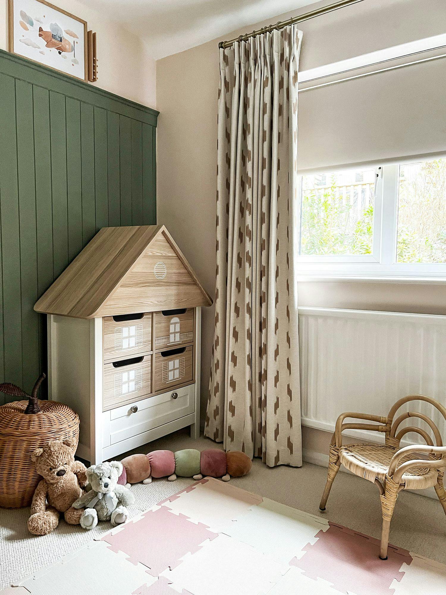 A wooden dollhouse shelf stands in a child's room with teddy bears on the floor, patterned curtains beside a window, and a small wicker chair, creating a cozy play area.