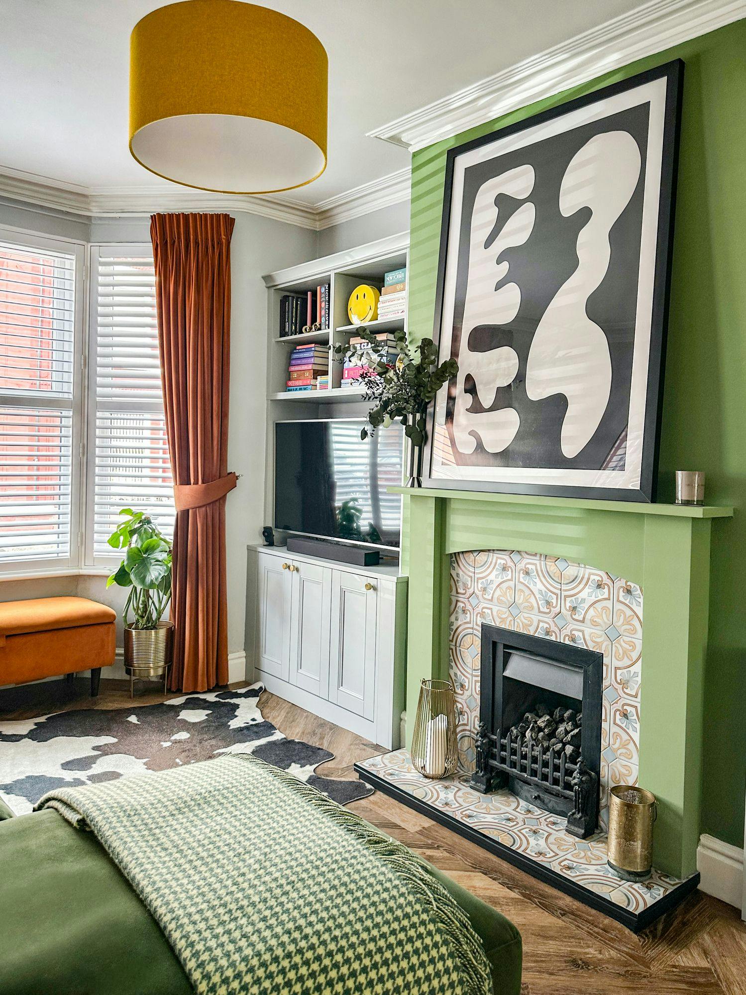 A modern living room features a large abstract artwork above a green-tiled fireplace. Beside it, shelves hold books and decor. An orange bench, green plant, and cowhide rug complete the space.