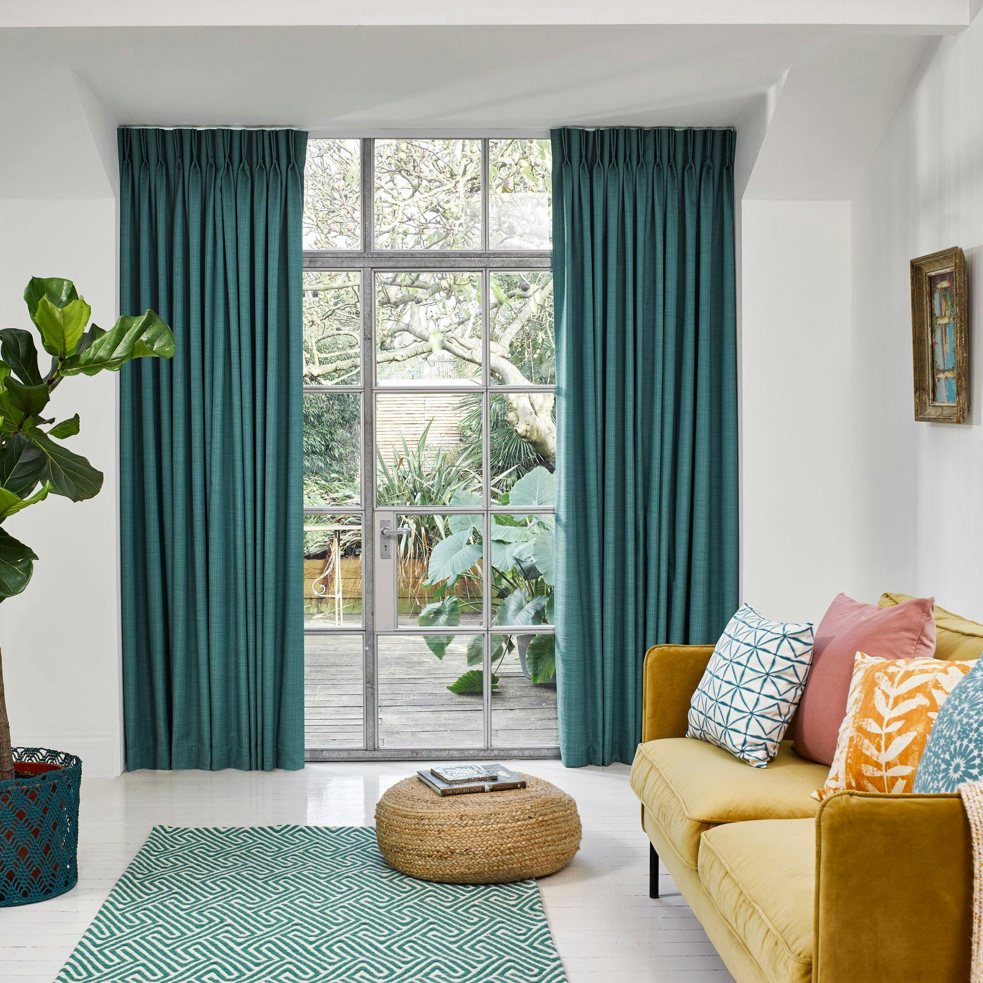 Teal curtains frame a large window, showcasing a garden. A mustard sofa with colorful cushions sits nearby, complemented by a wicker pouf, patterned rug, and a potted plant in a light, airy room.