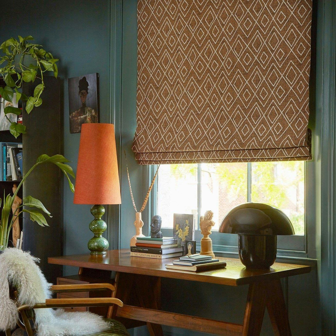 A wooden desk holds a green lamp, books, and decorative objects beneath a window. The brown Roman shade has a diamond pattern. Surrounding are green walls, plants, and a cozy chair.
