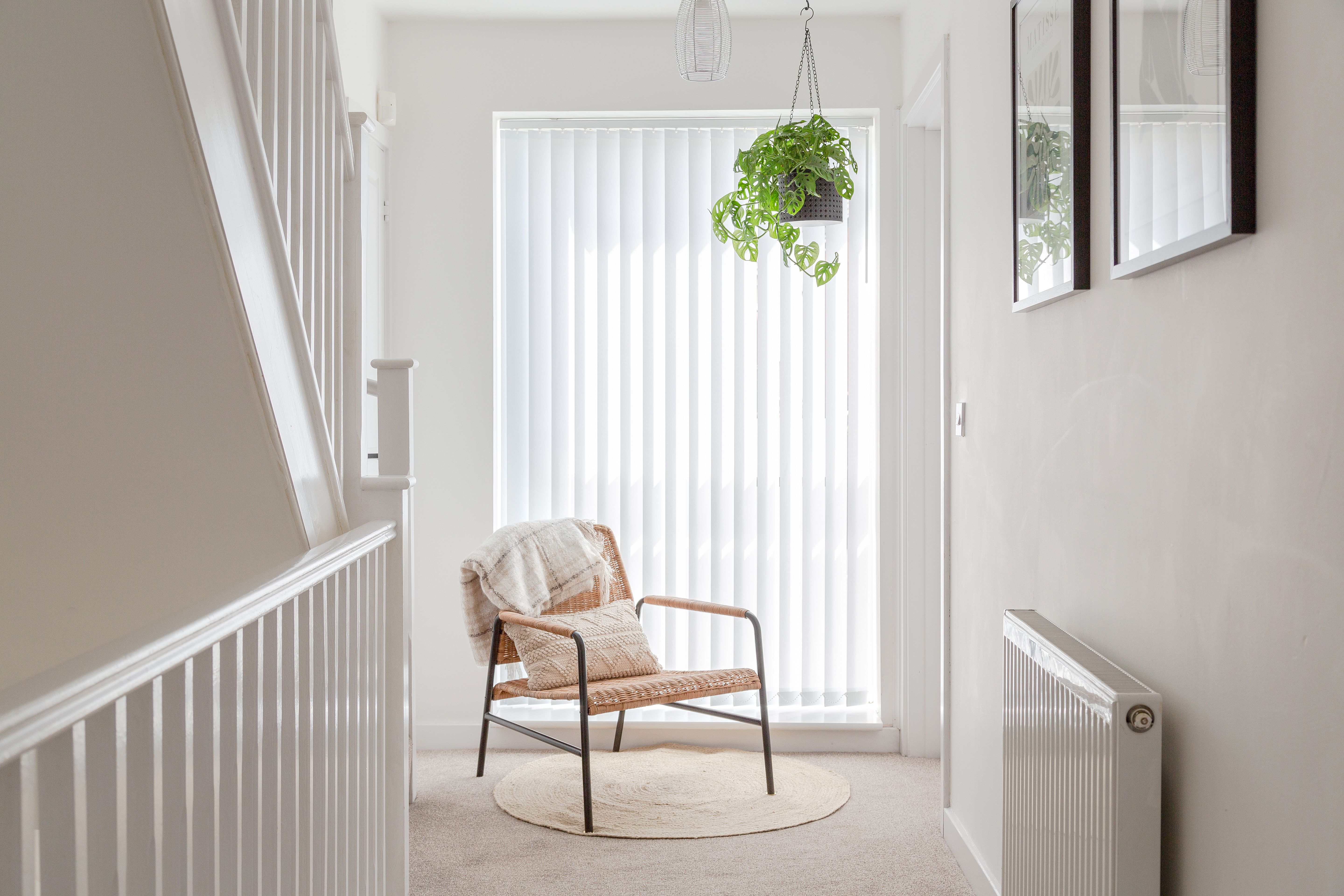 Chair sits calmly with a blanket draped over the back, bathed in light from vertical blinds. Above, a hanging plant adds greenery, surrounded by simple, minimalist decor in a hallway.