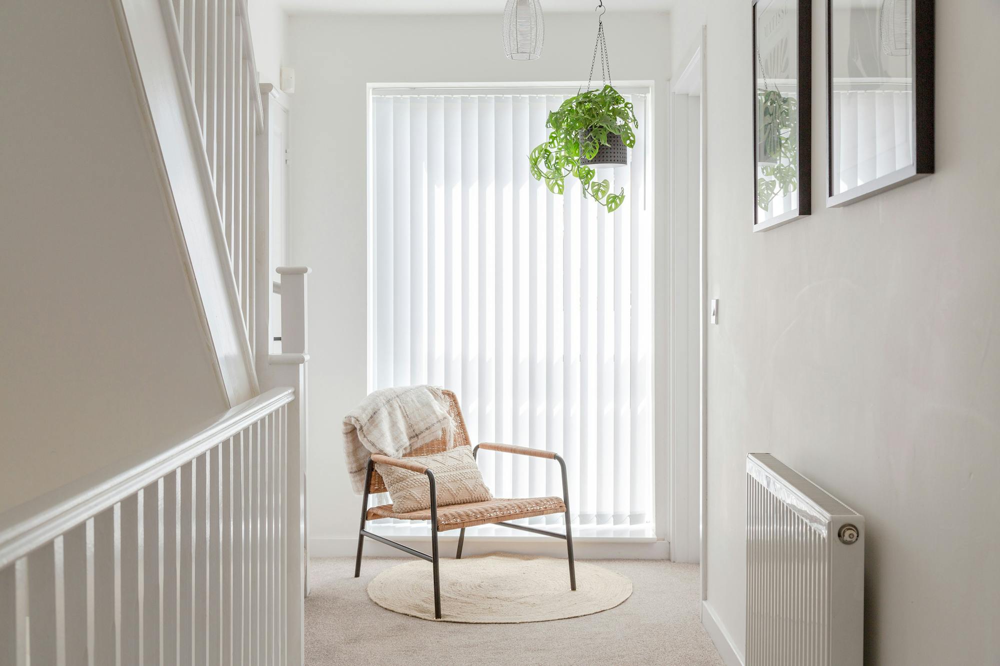 Chair sits calmly with a blanket draped over the back, bathed in light from vertical blinds. Above, a hanging plant adds greenery, surrounded by simple, minimalist decor in a hallway.